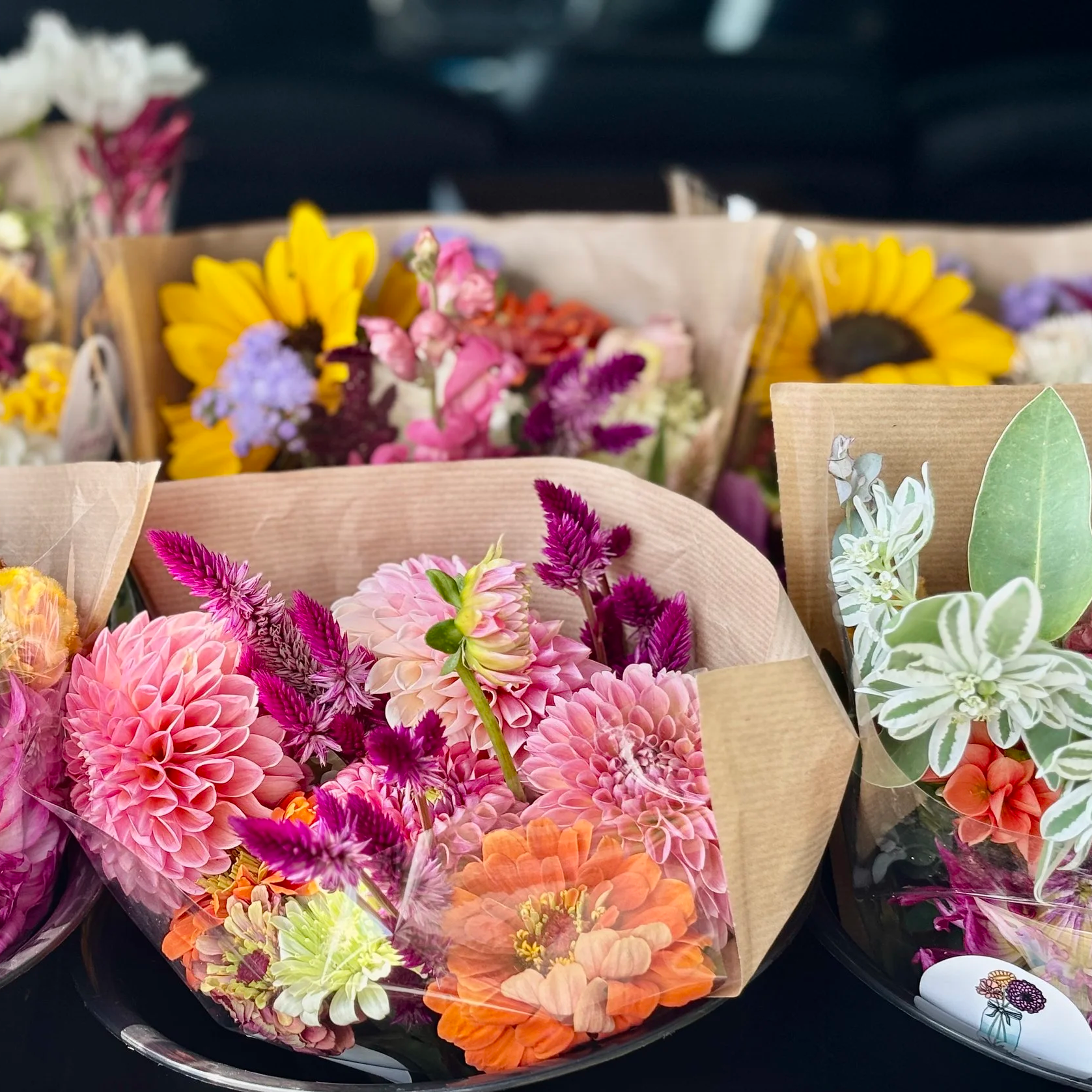 Colorful bouquets of flowers, including pink dahlias, orange zinnias, and purple celosia, wrapped in brown paper and plastic, displayed on a black surface.