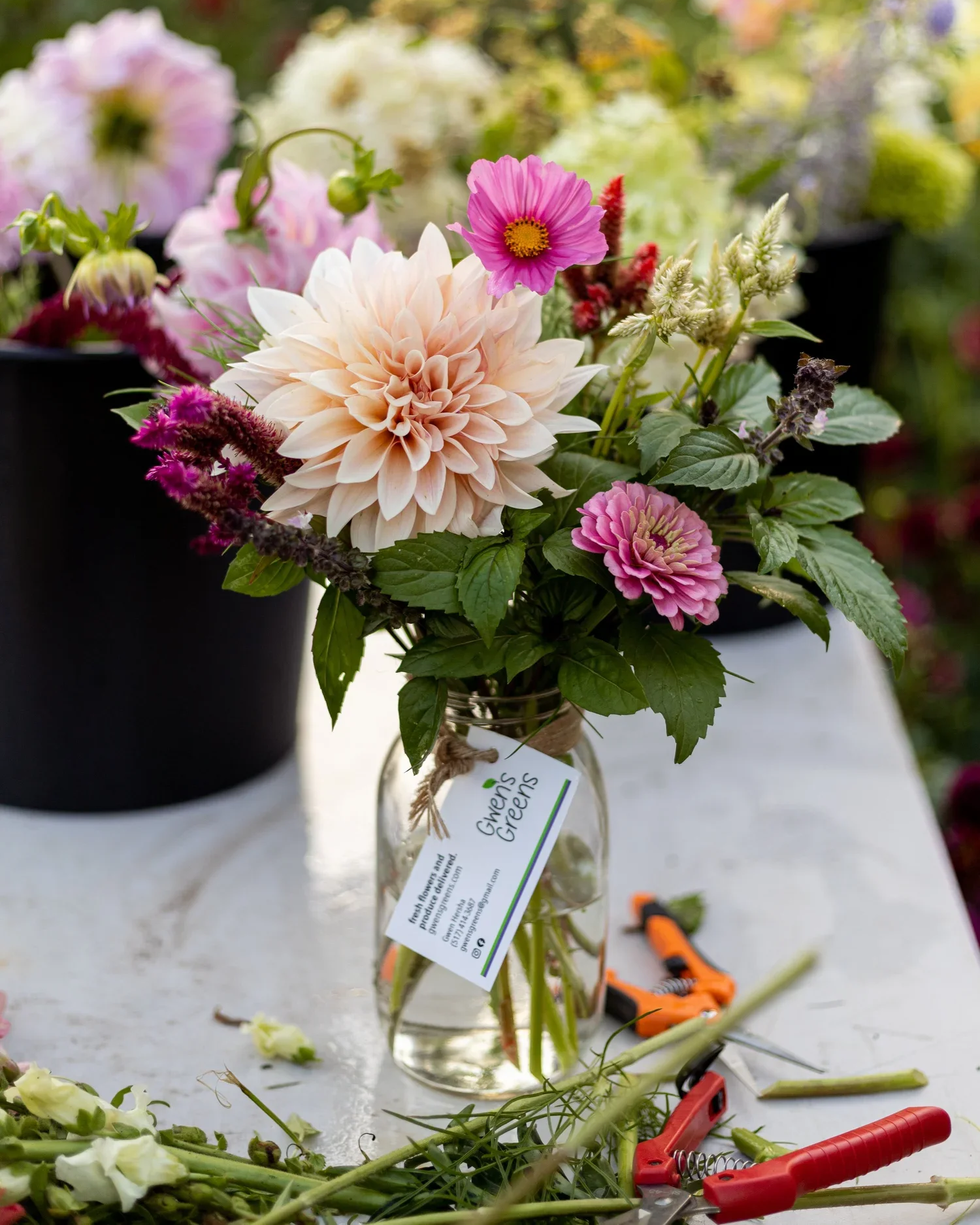 A glass jar with a floral arrangement of pink and white dahlias, pink cosmos, and other flowers, labeled 'Gwen's Greens', on a white table with gardening tools and plant cuttings nearby.