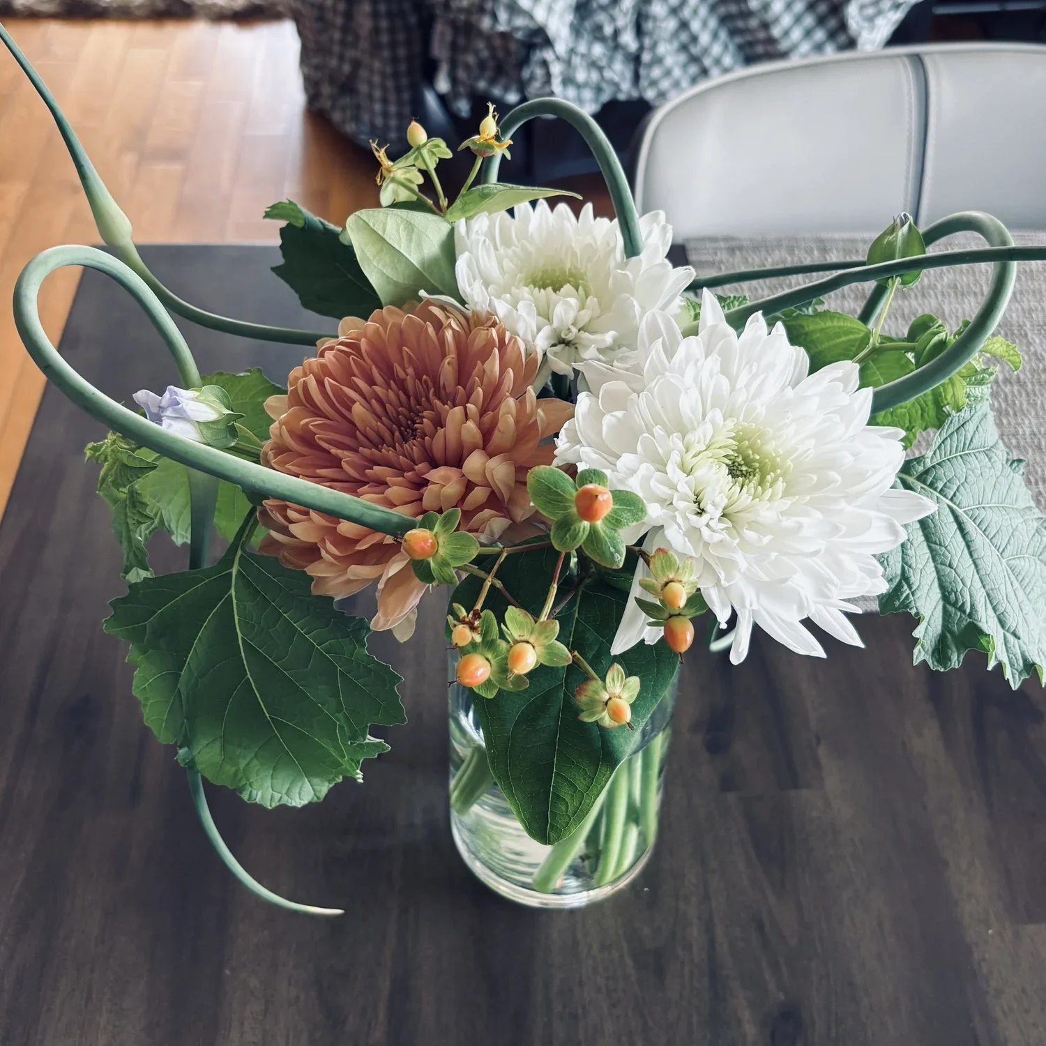 A vase of mixed flowers including white chrysanthemums, a brown dahlia, green leaves, and small orange berries on a dark wooden table.