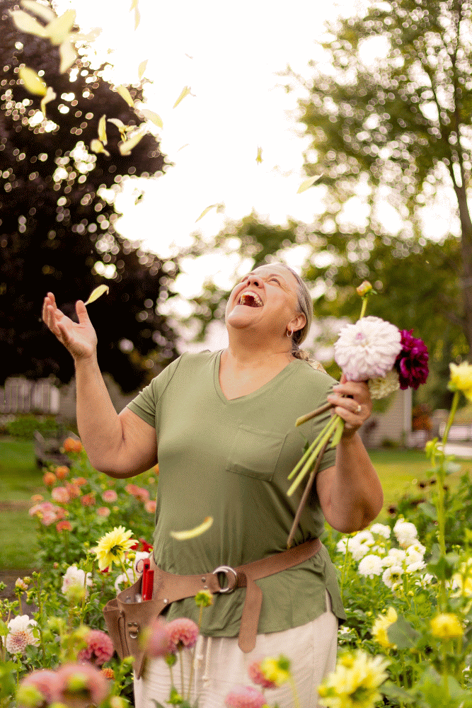 A woman laughing and throwing flower petals in the air while holding a bouquet of flowers in a garden.