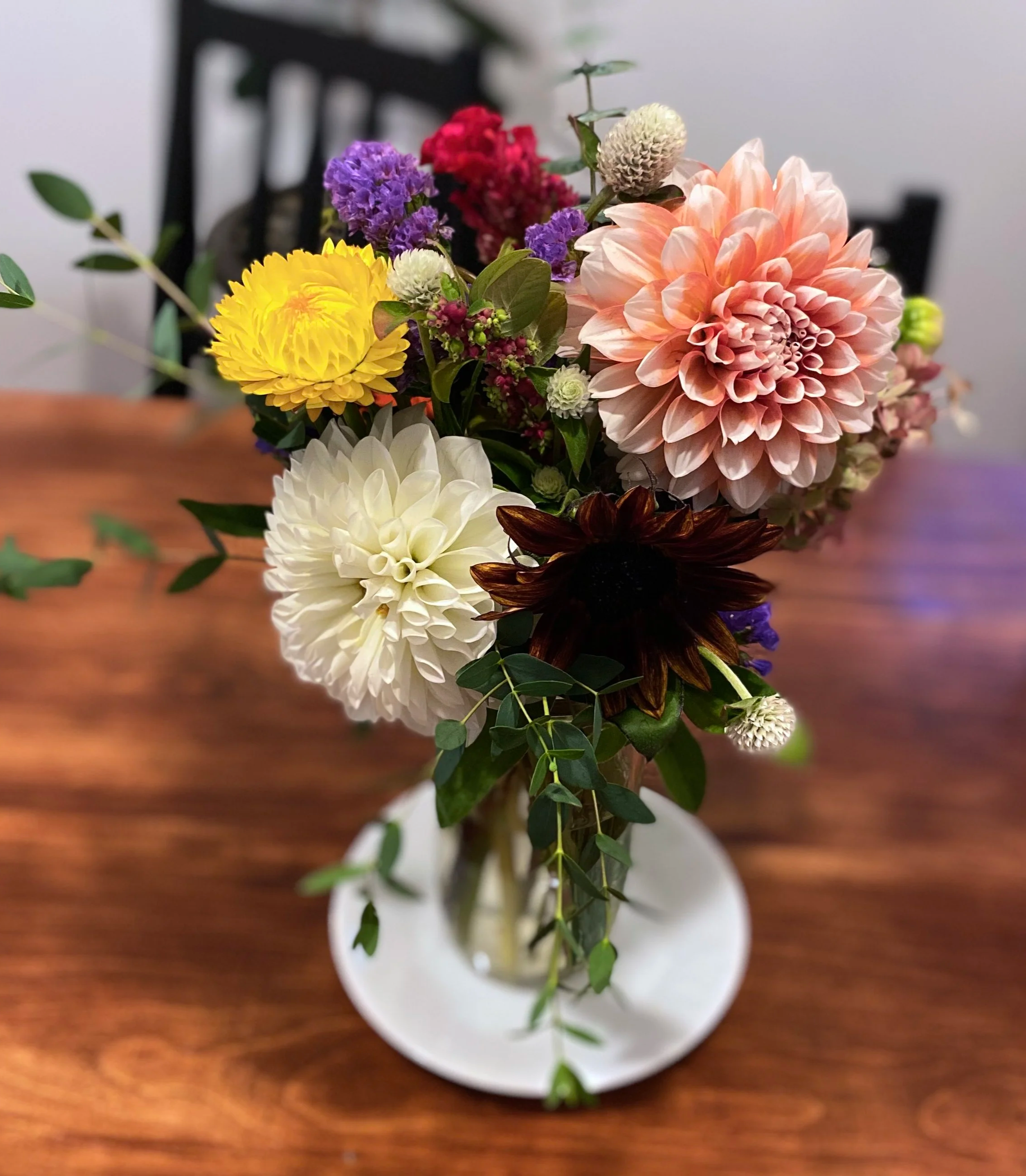 Colorful floral bouquet with dahlias, sunflowers, and sprigs of greenery in a white vase on a wooden table.