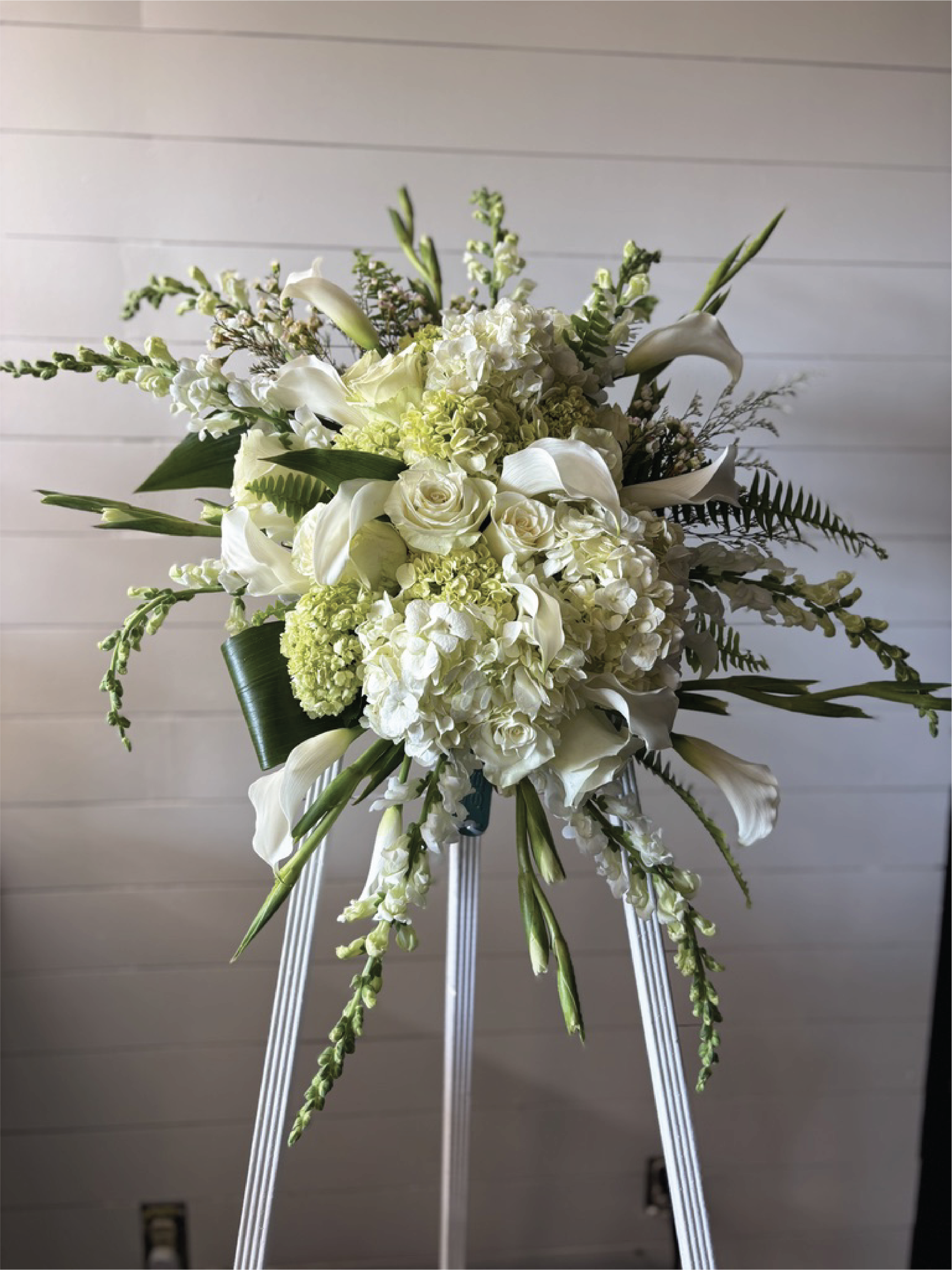 White floral arrangement on a stand with white roses, calla lilies, hydrangeas, and greenery against a light wood-paneled background.