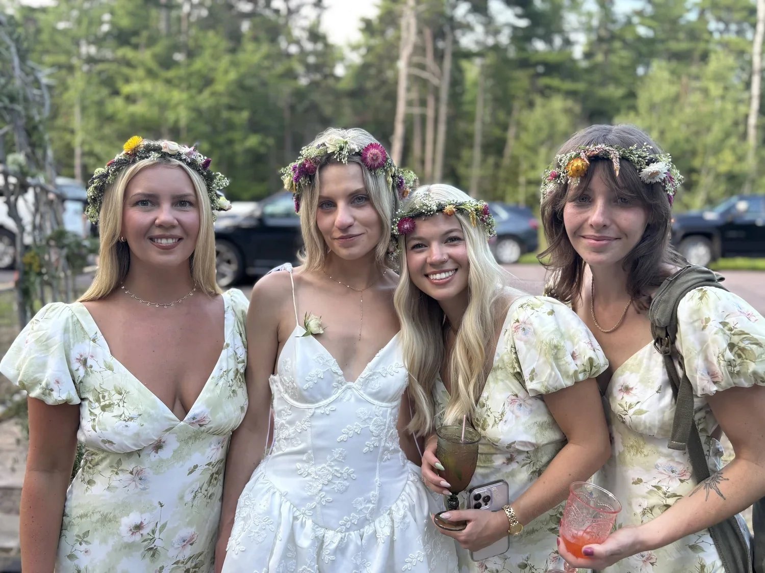 Four women celebrate a wedding outdoors, wearing flower crowns and floral dresses, with two holding drinks and one holding a phone.