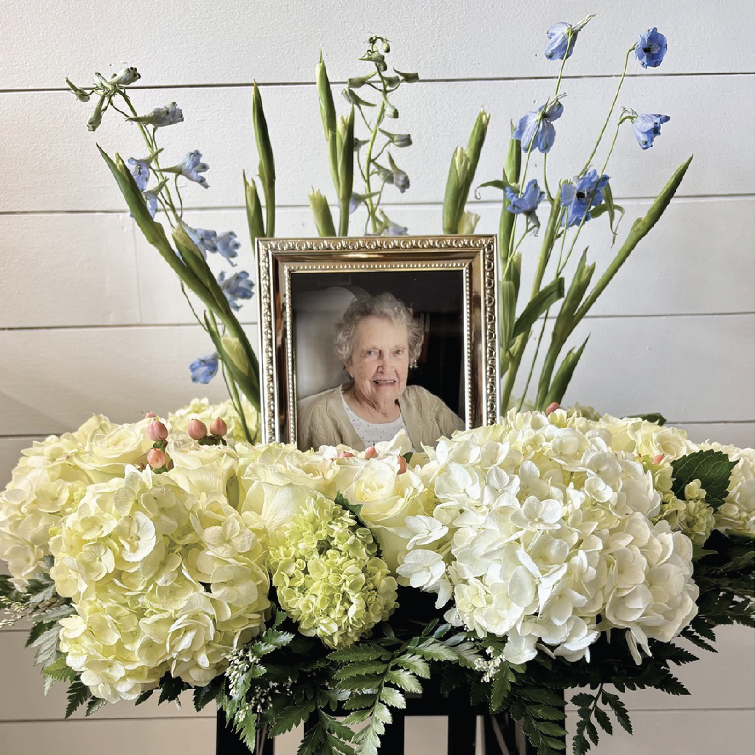 A framed photo of an elderly woman surrounded by a flower arrangement of white hydrangeas, roses, blue delphiniums, and green foliage, set against a white paneled wall.