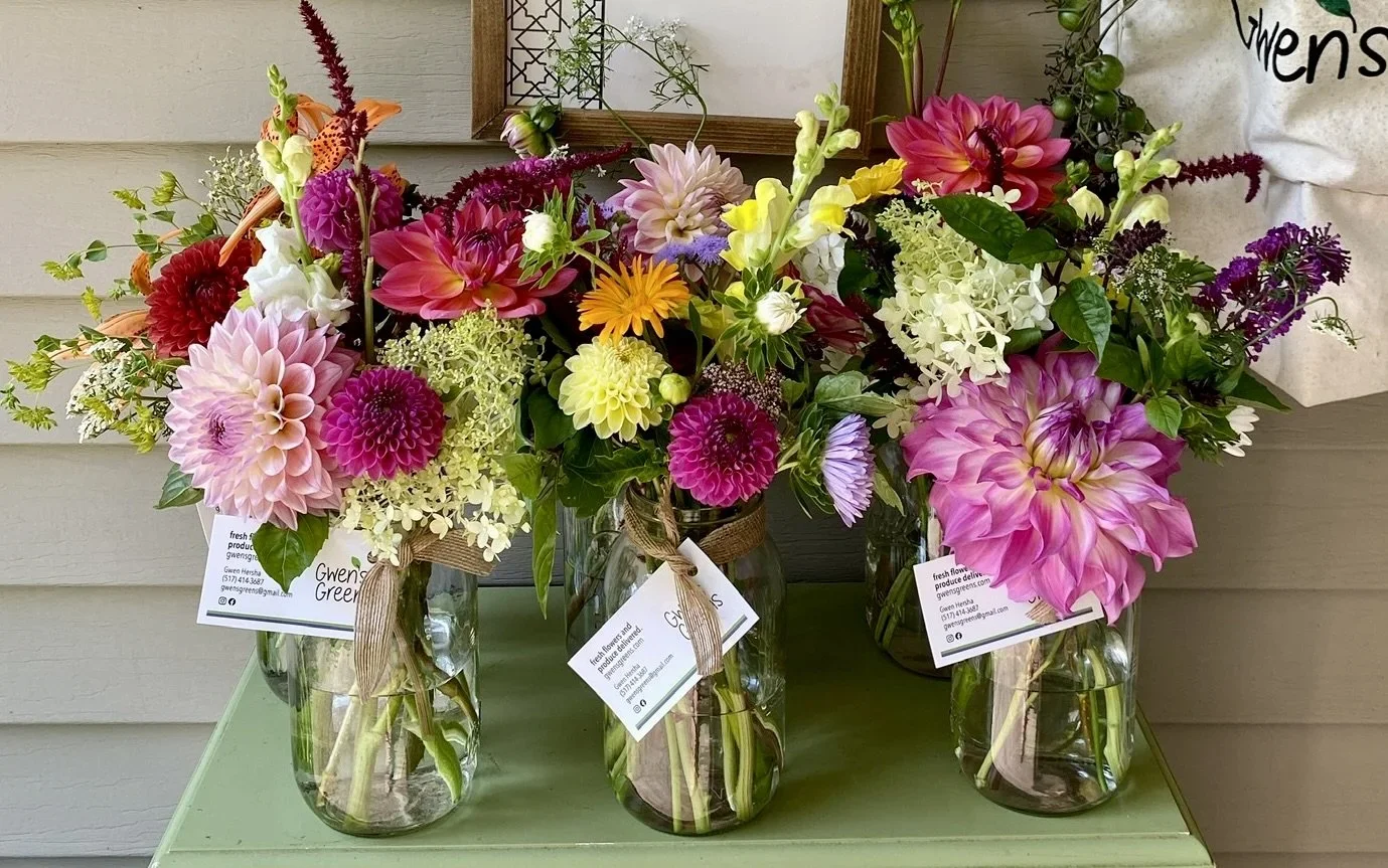 Three glass jars filled with colorful fresh flower bouquets including dahlias, zinnias, and lilies placed on a light green surface against a wall with a framed display.