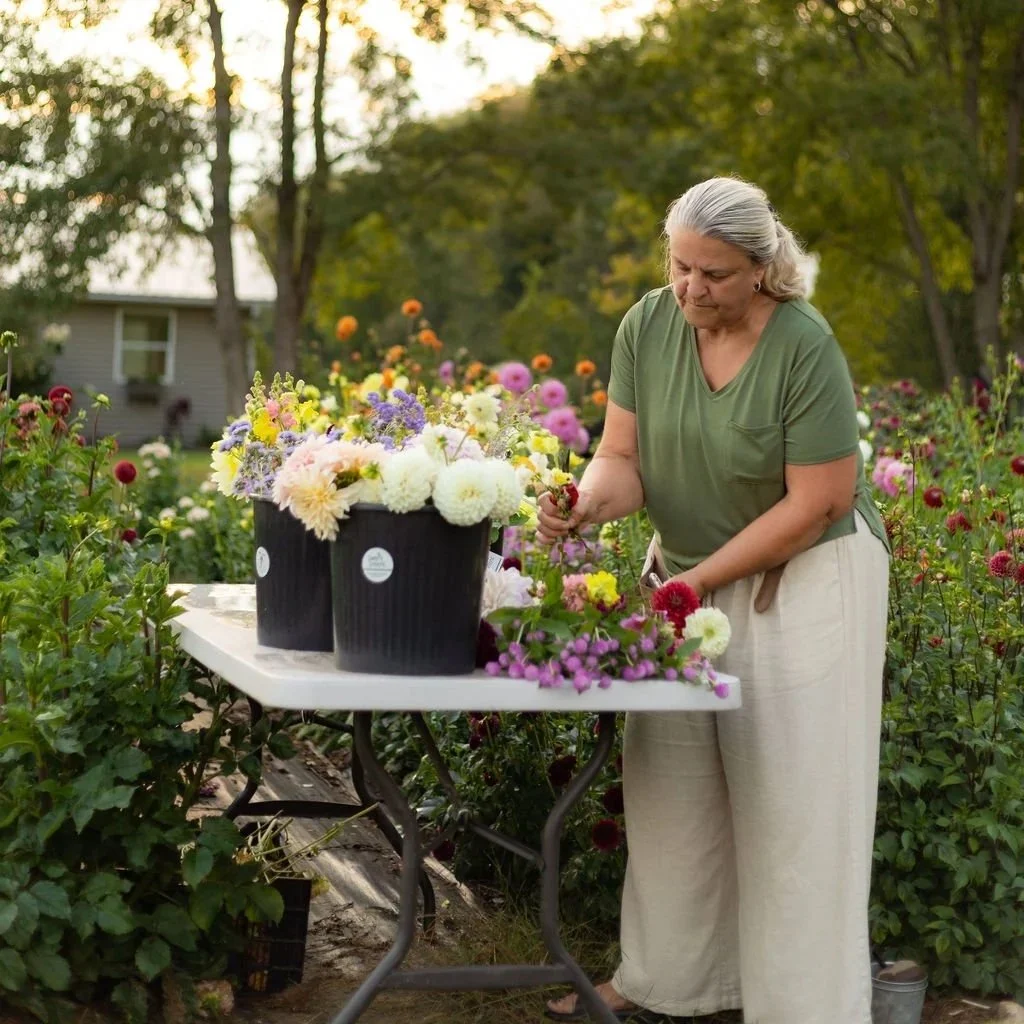 An elderly woman arranging colorful flowers on a white table outdoors, surrounded by a garden in bloom.