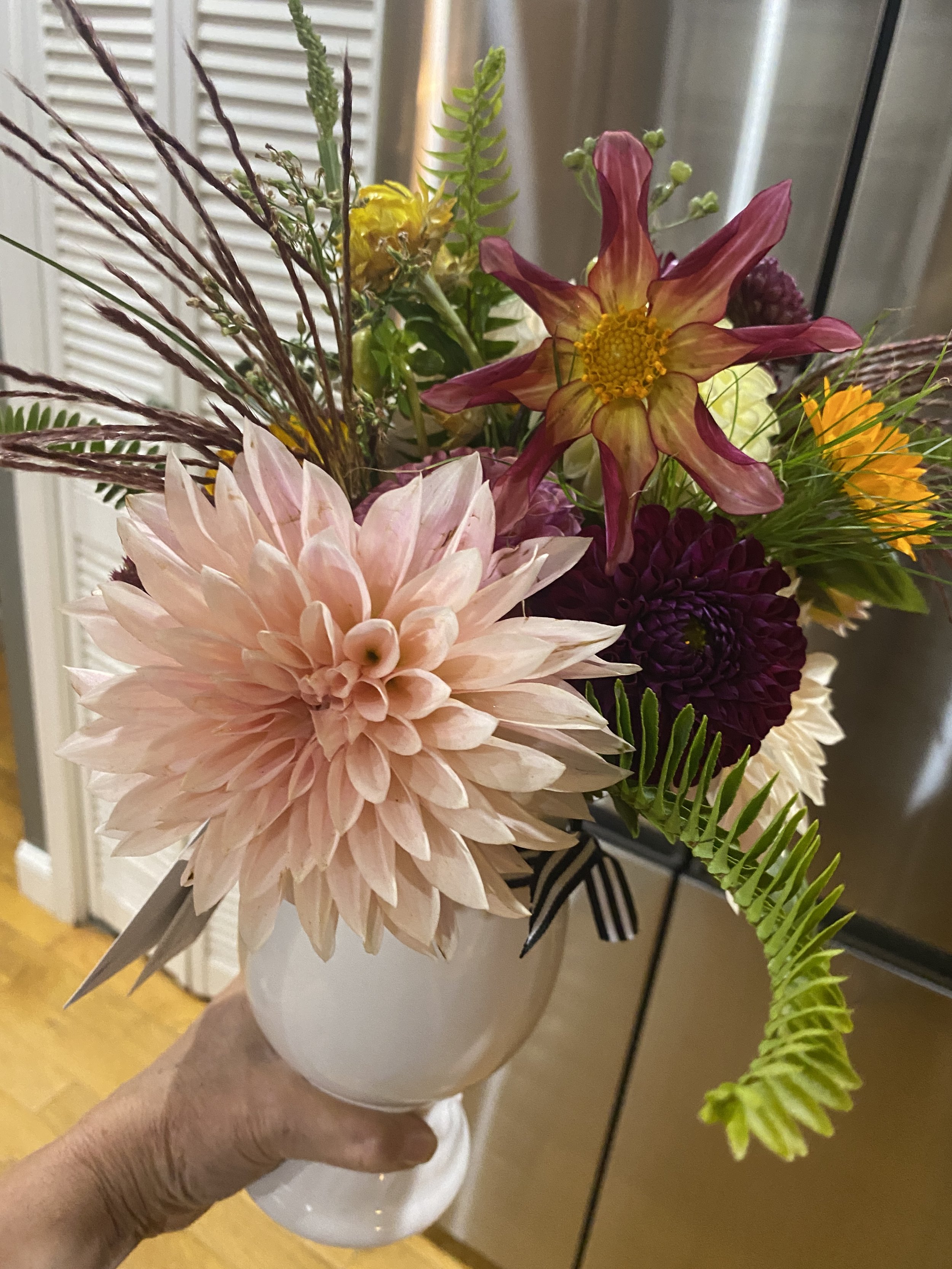 A hand holding a white vase with a colorful bouquet of various flowers, including a large peach dahlia, pink and yellow dahlia, burgundy, purple, and orange flowers, with green fern leaves and decorative grasses.