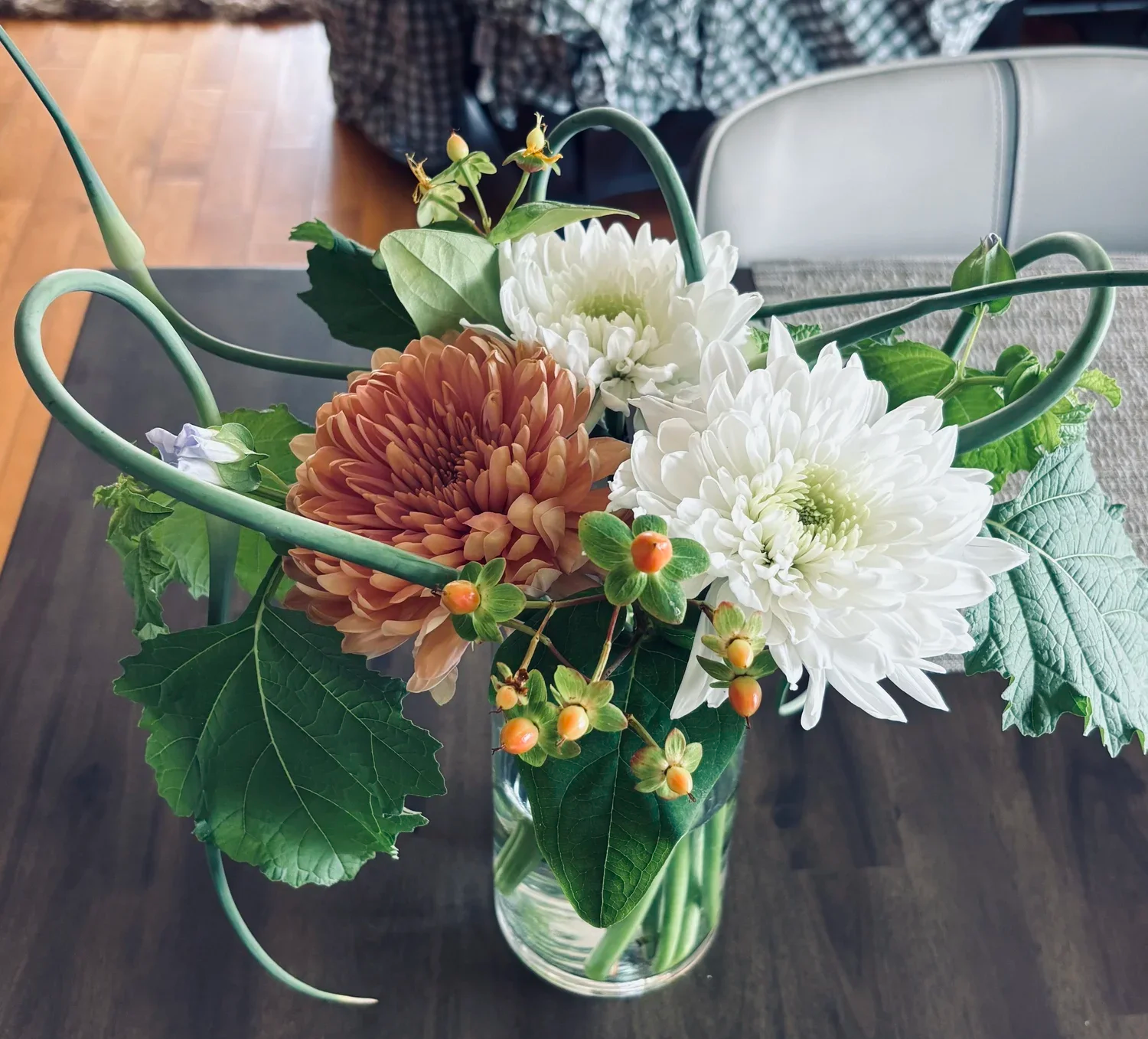 A floral arrangement in a glass vase with white and orange dahlias, green leaves, and small berries, set on a dark wooden table.