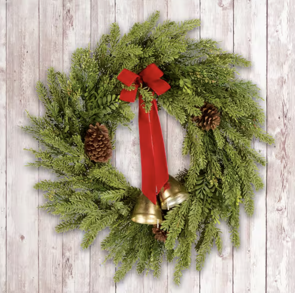 Christmas wreath made of green pine branches, decorated with pine cones, a red ribbon bow, and two gold bells, hanging on a white wooden background.