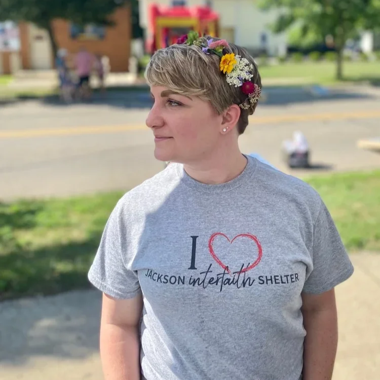 A woman with short blonde hair wearing a light grey T-shirt that says "I love Jackson interfaith shelter" with a red heart. She has a floral headband and is looking to the left. The background is a park or outdoor area with trees, grass, and a few blurred people.
