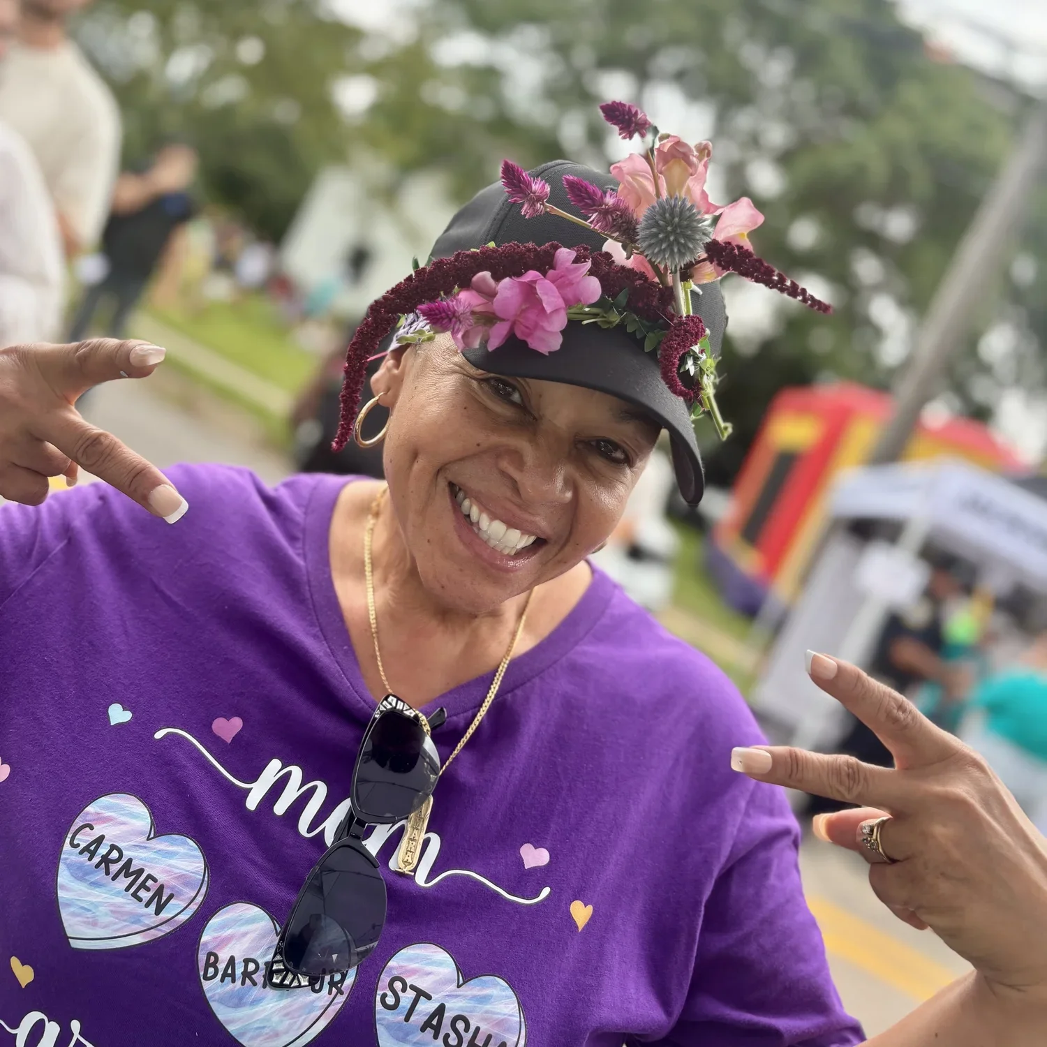 A woman smiling and pointing at her purple T-shirt, wearing a floral hat, at an outdoor event.
