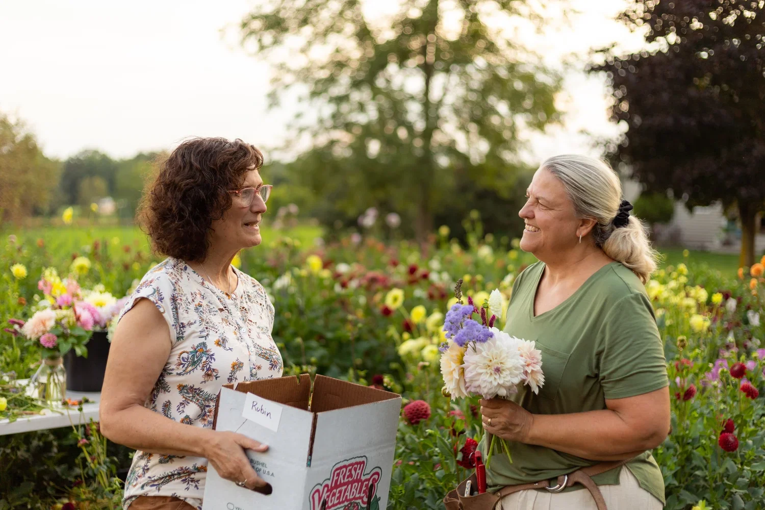 Two women smiling and talking in a garden filled with colorful flowers, one holding a box labeled 'Robin' and the other holding a bouquet of flowers.