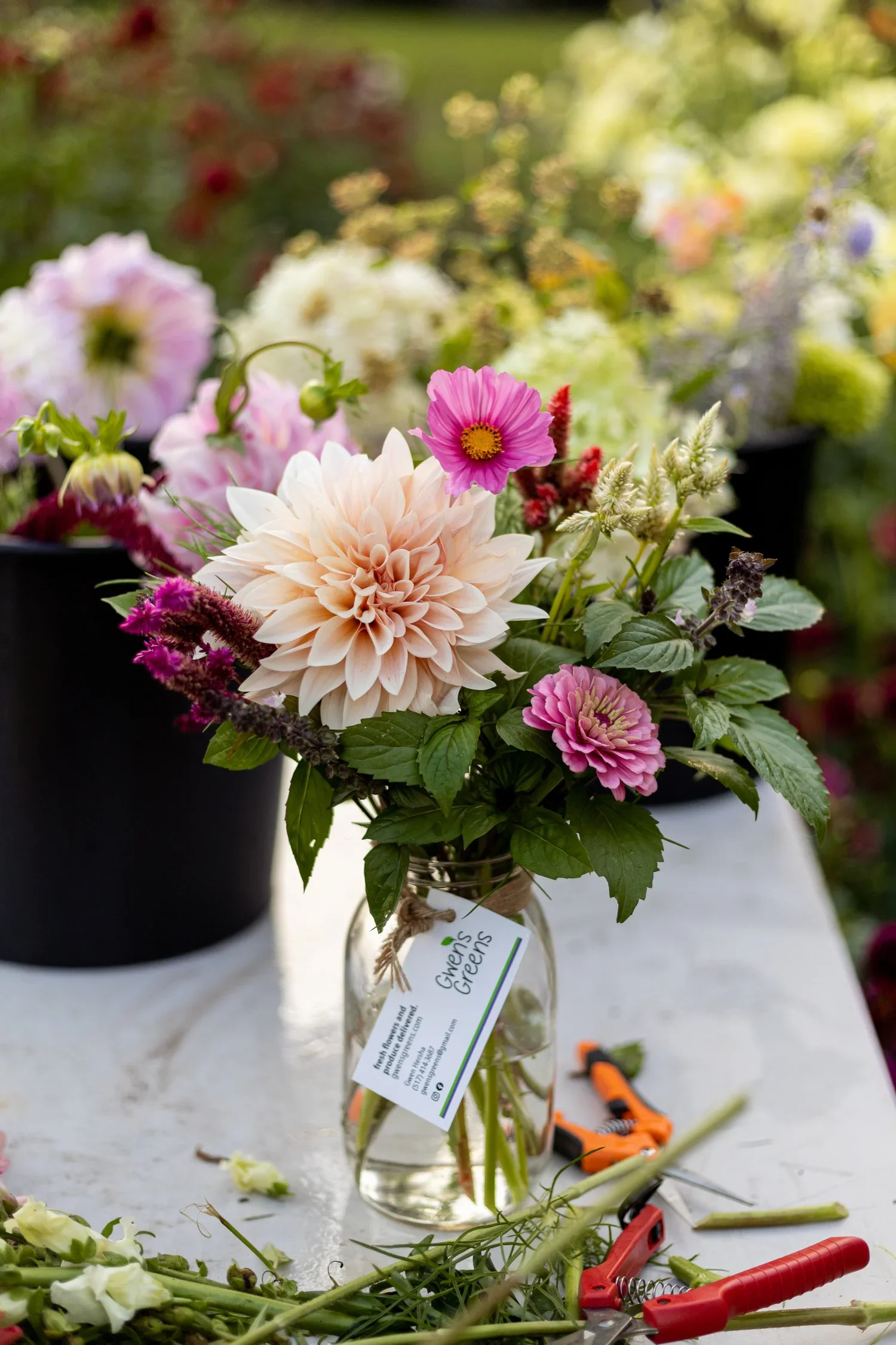 A glass jar with a bouquet of pink, white, and purple flowers on a white table, surrounded by gardening tools and plant cuttings, with a blurred background of greenery and plants.