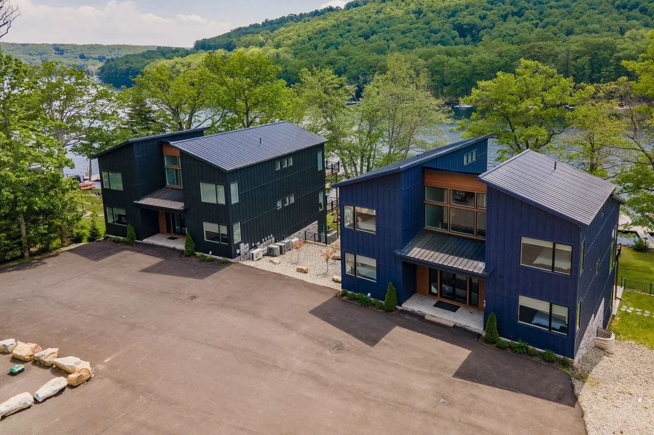 Two modern houses with dark blue and black exteriors are situated near a lake with green trees in the background.