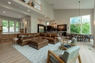 Spacious living room with large sectional sofa, armchair, dining table, and windows showing greenery outside.
