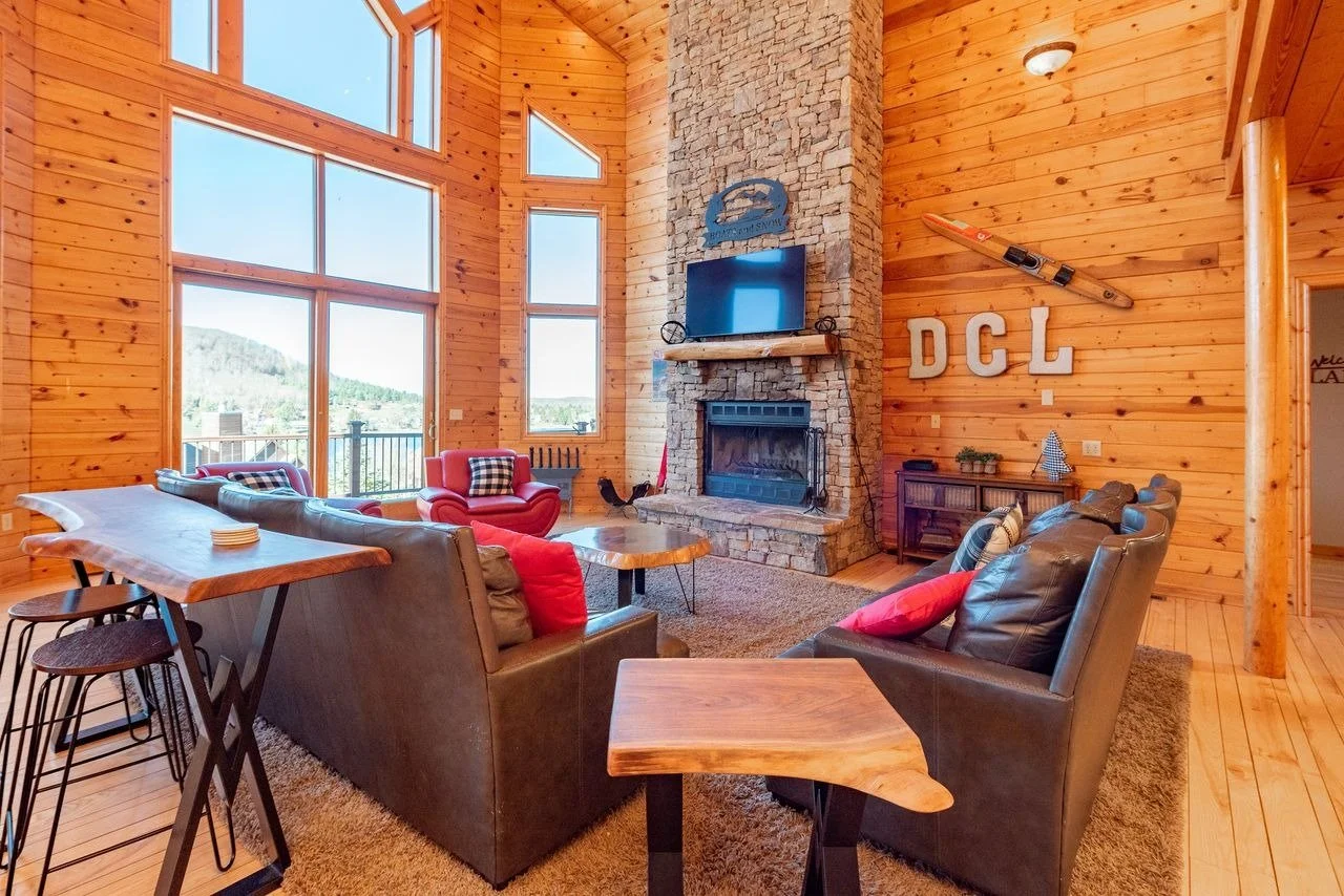 Living room with high wooden walls and large windows showing mountain views, featuring a stone fireplace with a mounted TV, leather and fabric chairs, a wooden coffee table, and decorative items including ladder and letters on the wall.