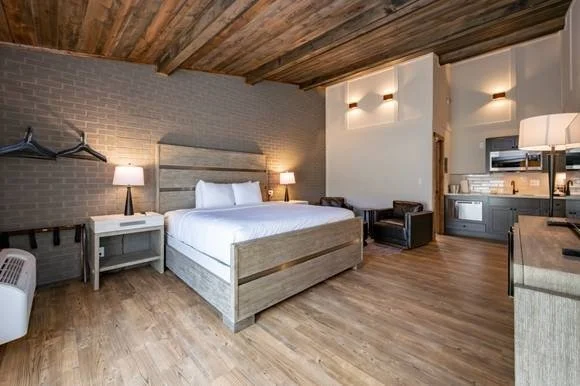 Modern hotel room with a wooden ceiling, brick wall, large bed with white linens, two nightstands with lamps, a small sitting area, and a kitchenette.