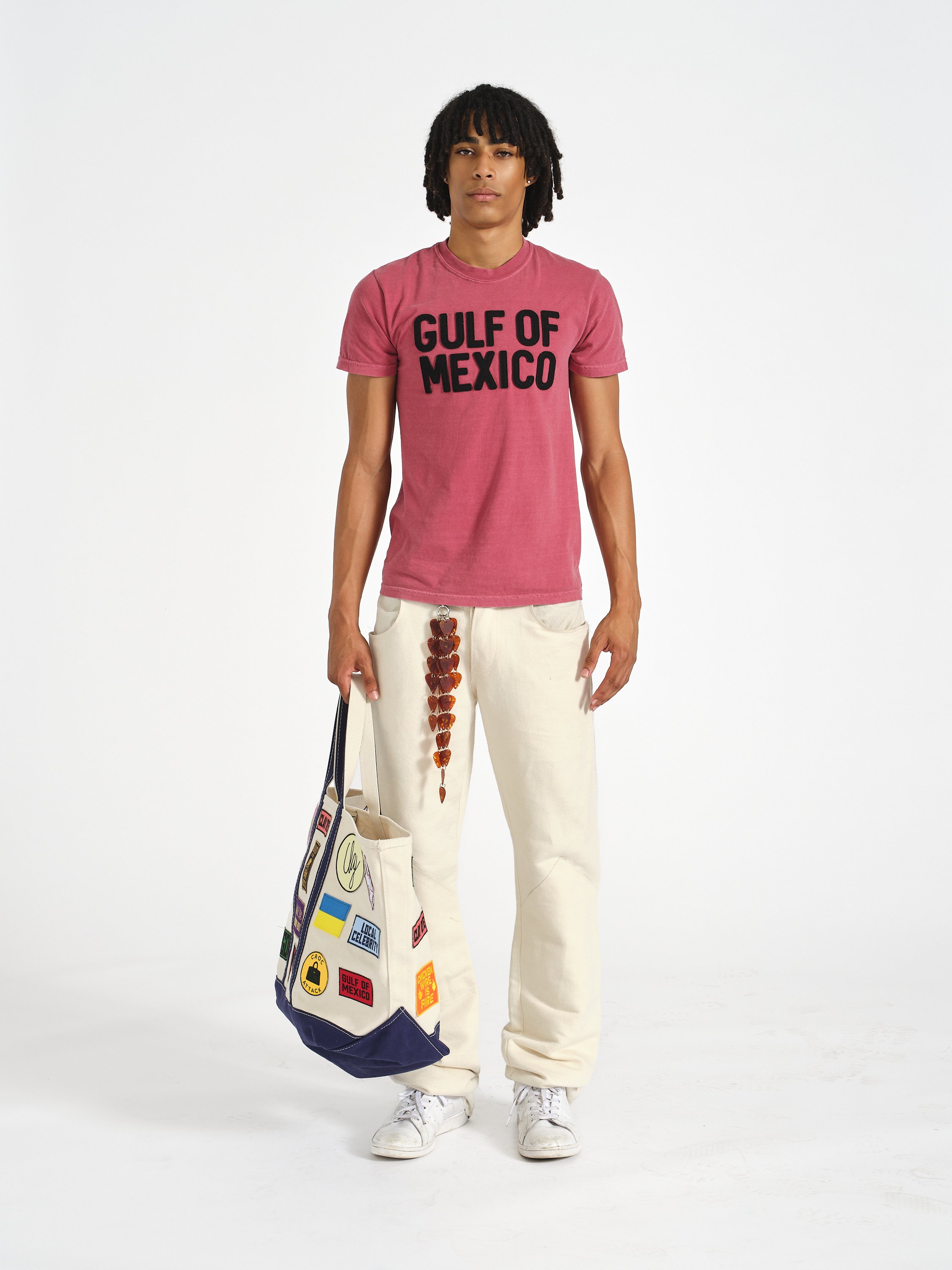 A male model stands in Clay Dye denim holding an extra large tote bag full of patches. He wears a red shirt that says Gulf of Mexico.