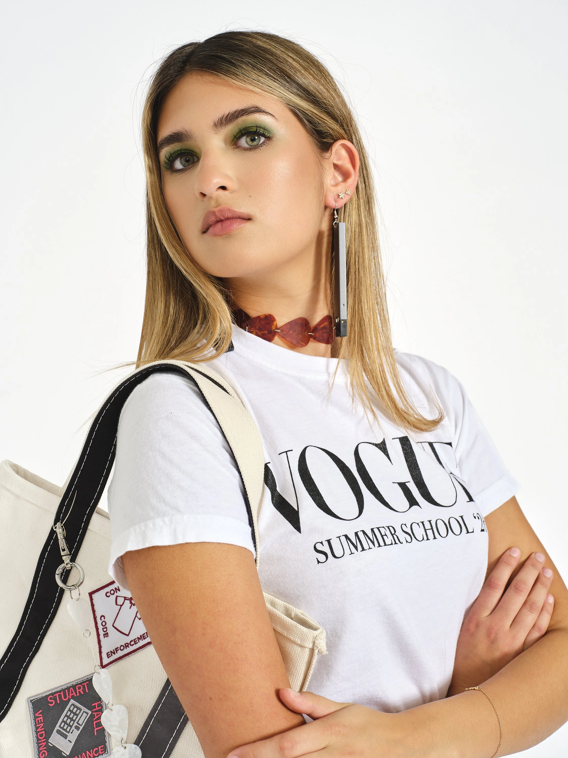Young woman with green eye makeup, long earrings, and a white “Vogue” summer school T-shirt posing against a white background