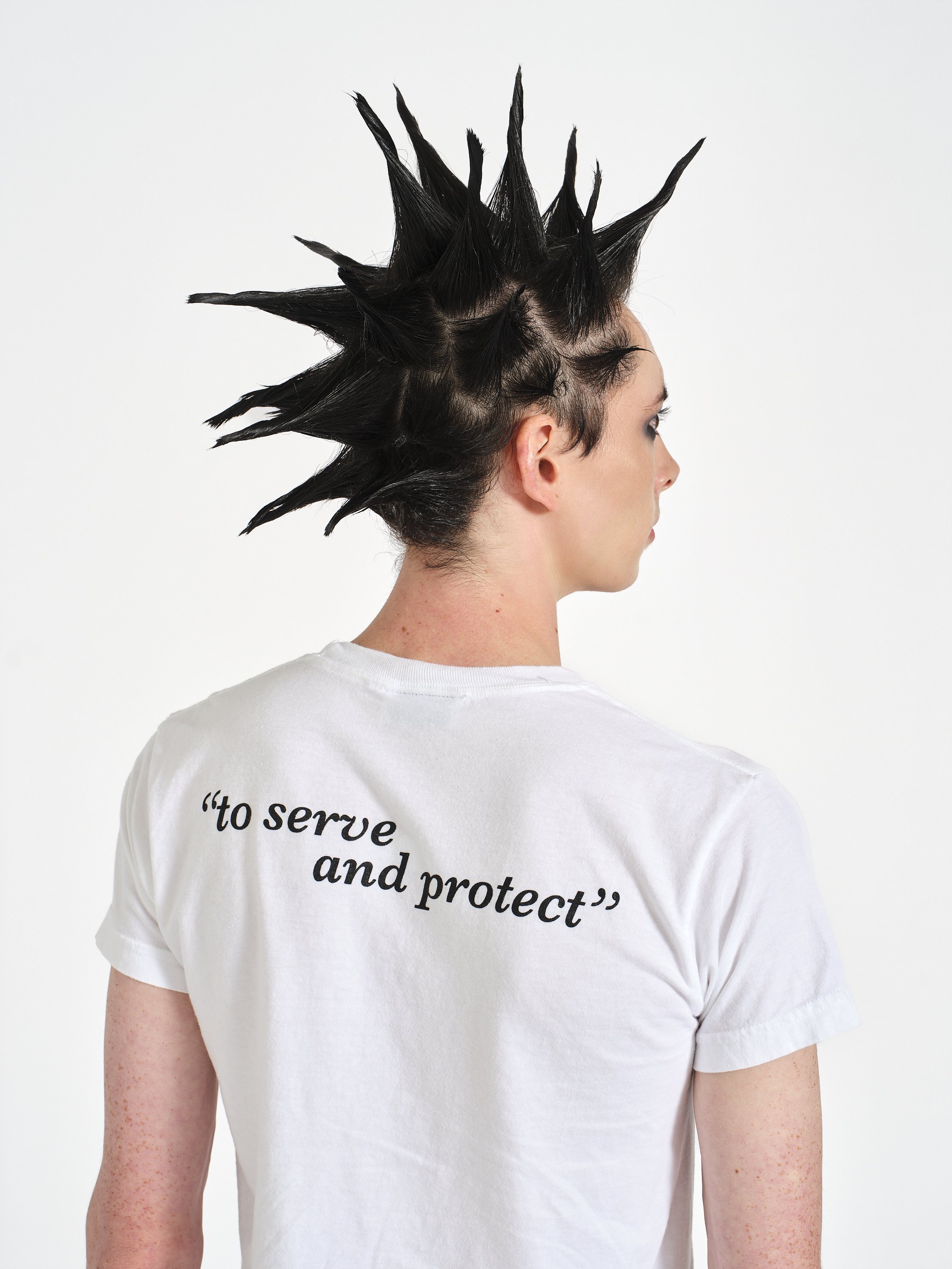 Person in profile with black spiked hair and visible scalp, wearing a white graphic T-shirt.