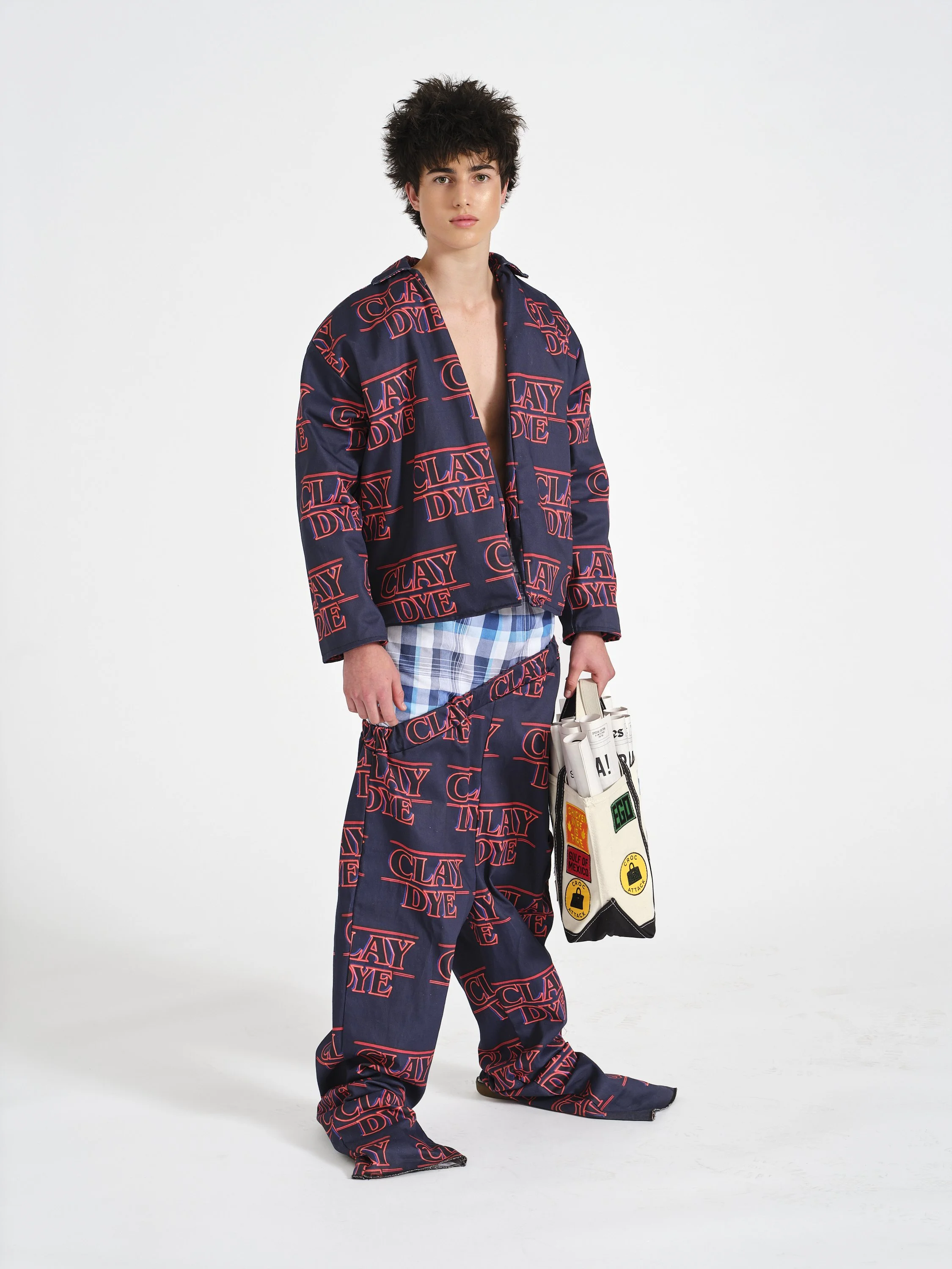 Male model wearing a navy blue suit with red 'CLAY DYE' logo in the Stranger Things font, holding a white tote bag with various patches and designs against a plain white background.