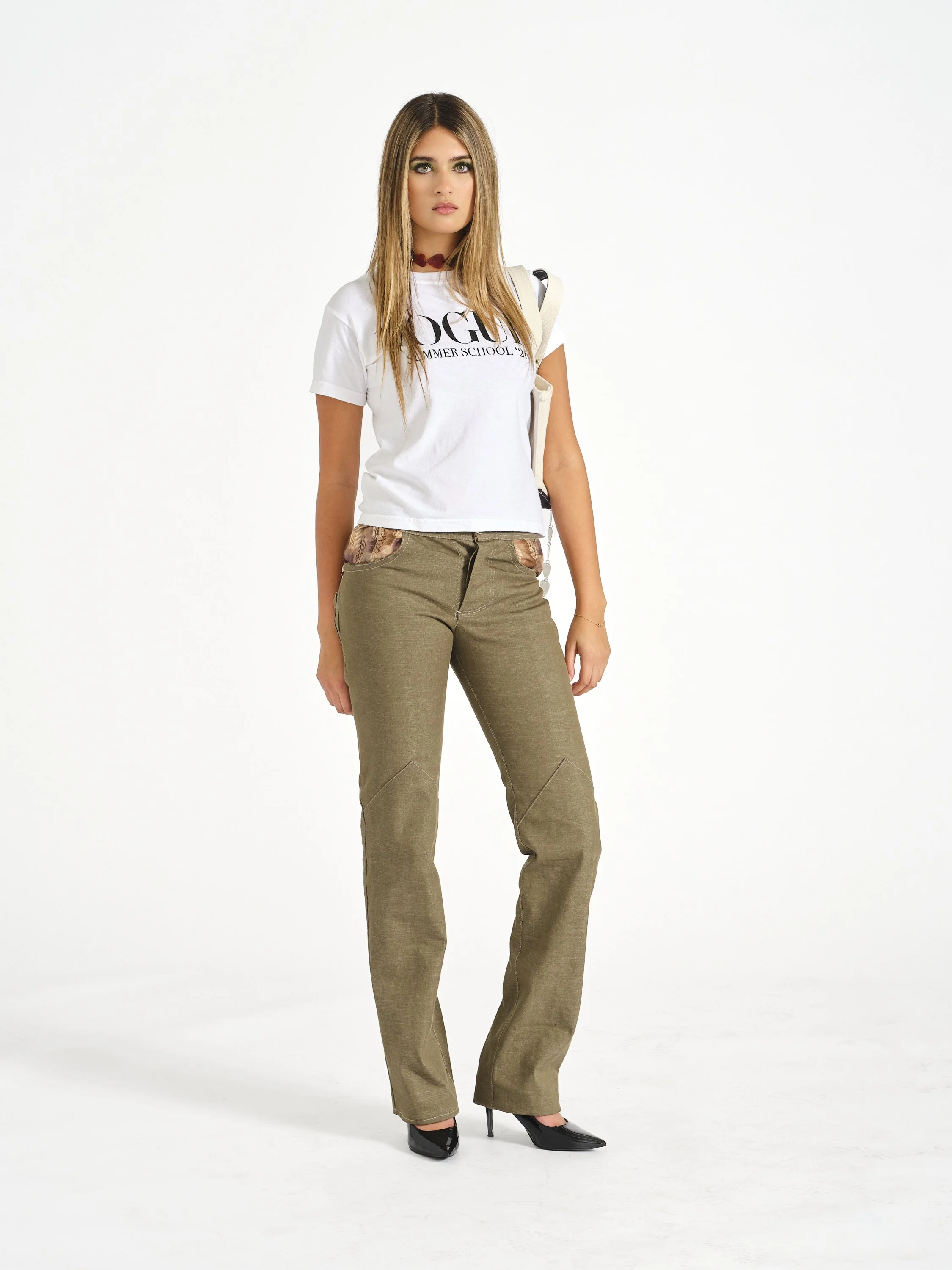 Female model with long blonde hair wearing a white T-shirt, khaki pants, black high heels, and a red choker necklace, standing against a plain white background.