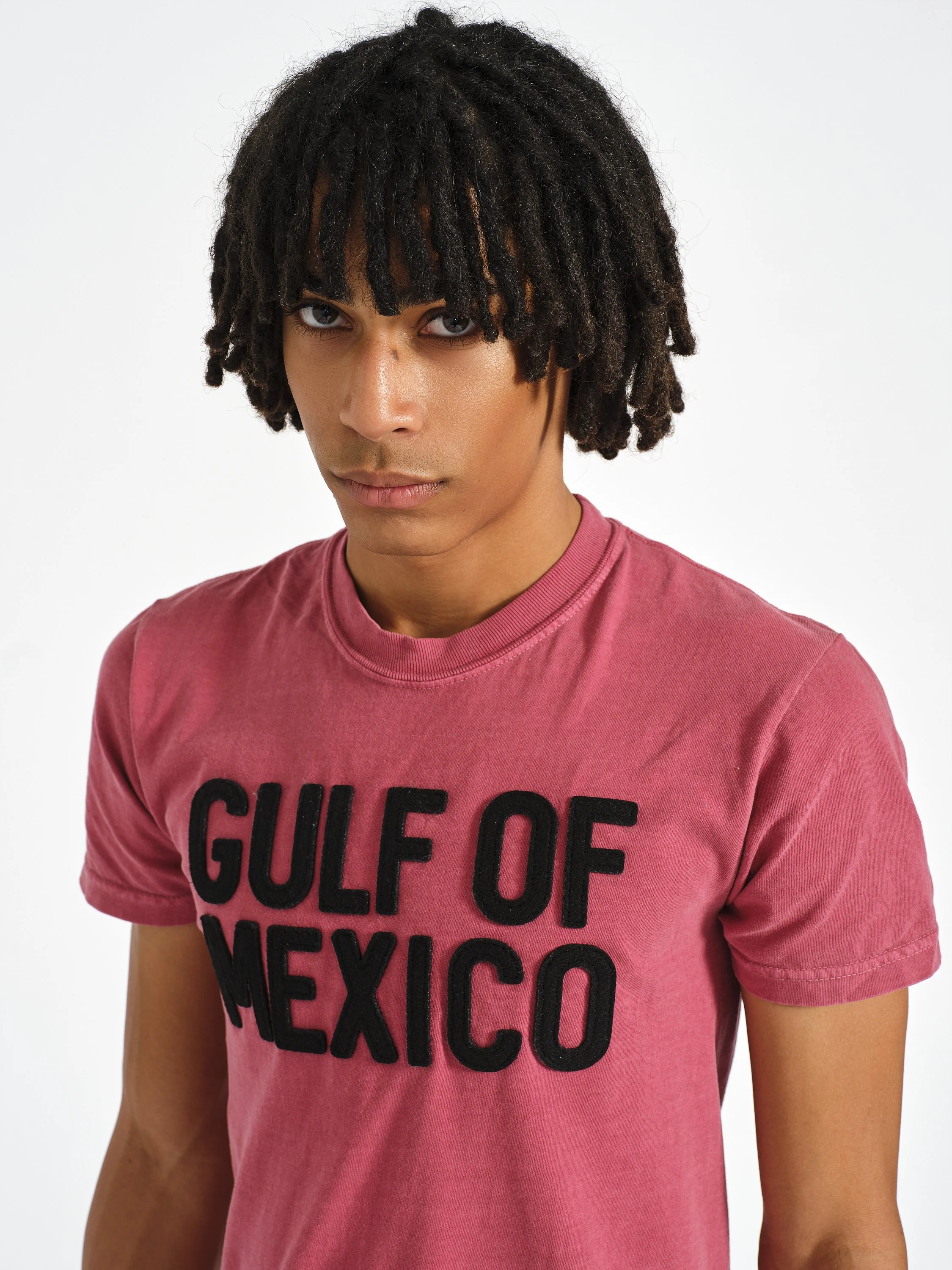 Close-up portrait of a person with curly black hair wearing a pink T-shirt reading “GULF OF MEXICO” against a white background