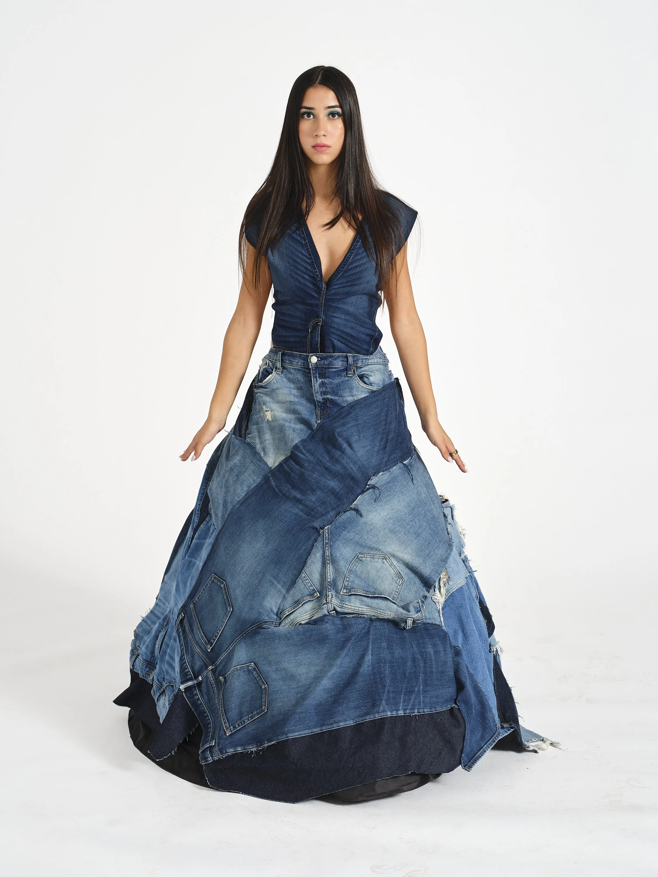 A woman with long dark hair wearing a denim vest and a skirt made from patched denim jeans, standing against a plain white background.