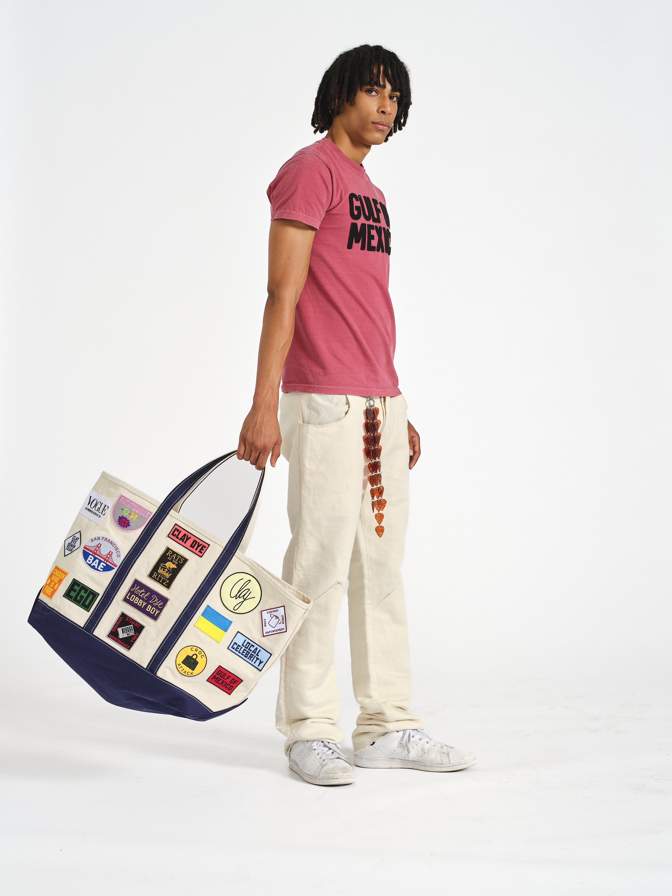 Fashion model wearing a pink graphic T-shirt, cream pants, and white sneakers while carrying a patch-covered canvas tote bag.