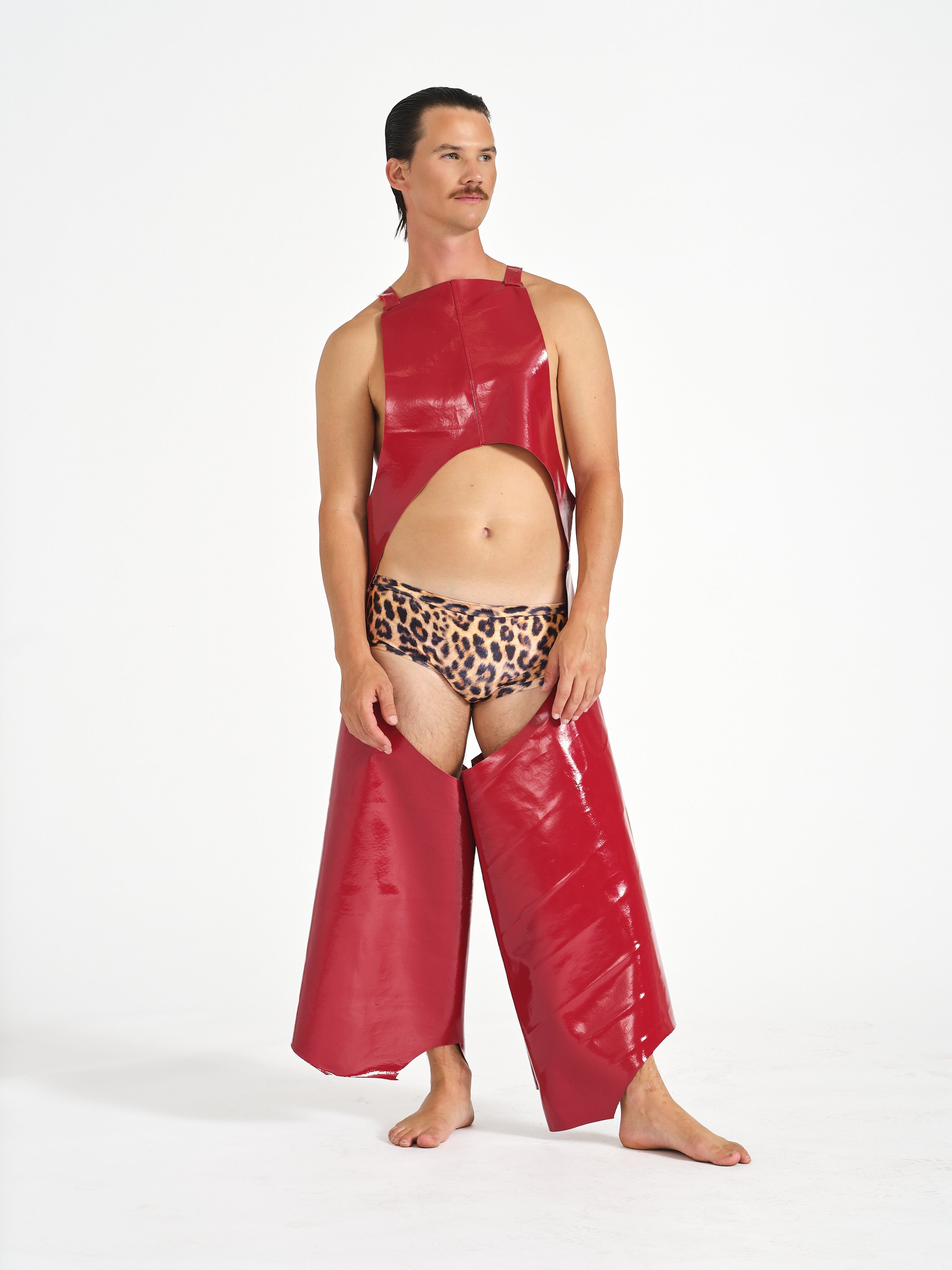 Male model standing barefoot in a pose, wearing leopard print underwear, and a pair of shiny red leather chaps, with a plain white background.