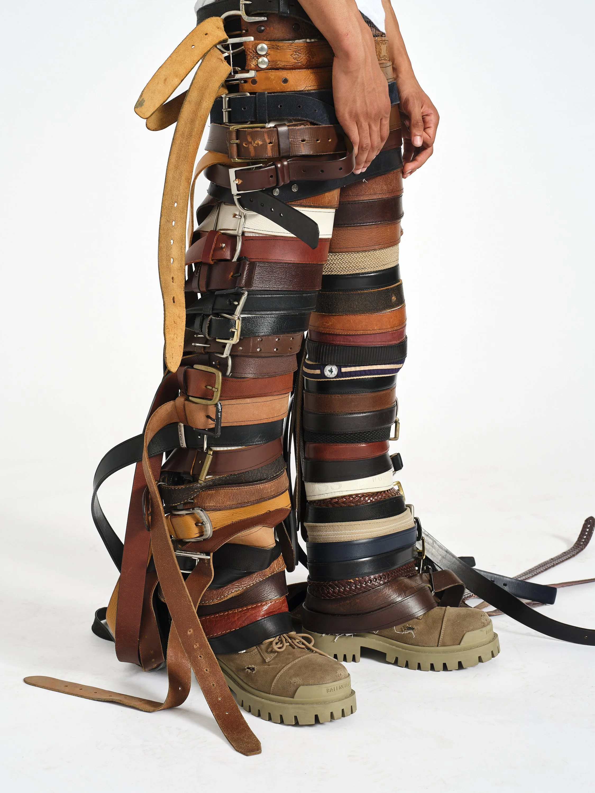 Person wearing multiple layered leather belts and tan work boots against a white background