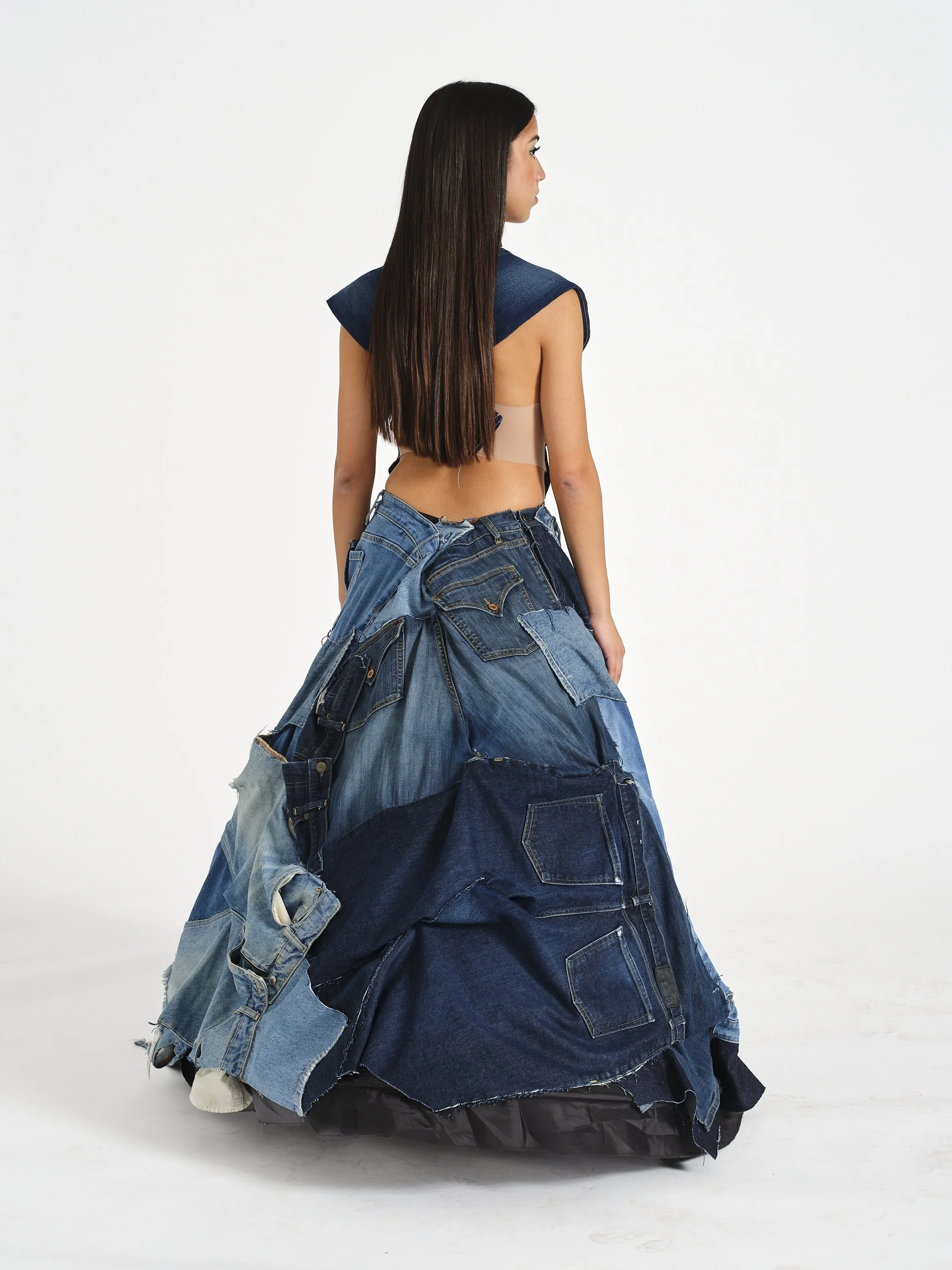 Woman with long dark hair wearing a blue top and oversized patchwork denim skirt, shown from behind against a white background