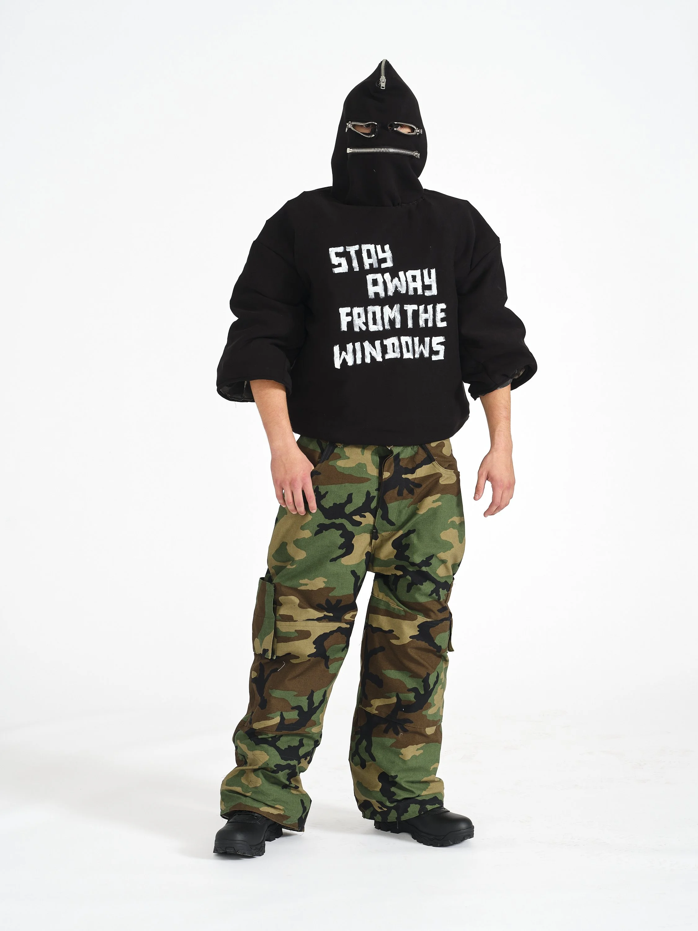 A male model wearing a black hoodie with Zipper and eye holes, camouflage pants, and black boots, standing against a plain white background.