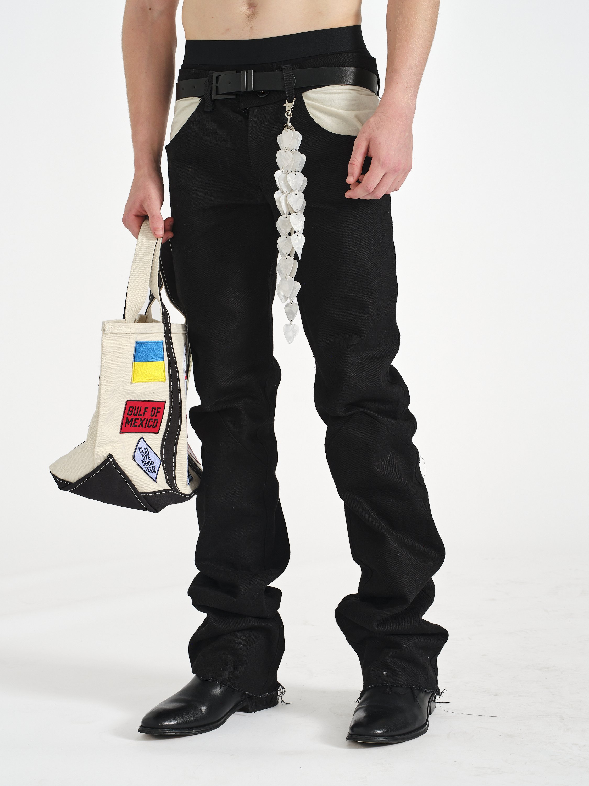 Person wearing black pants and boots, carrying a patched canvas tote bag and a heart-shaped white shell belt accessory.