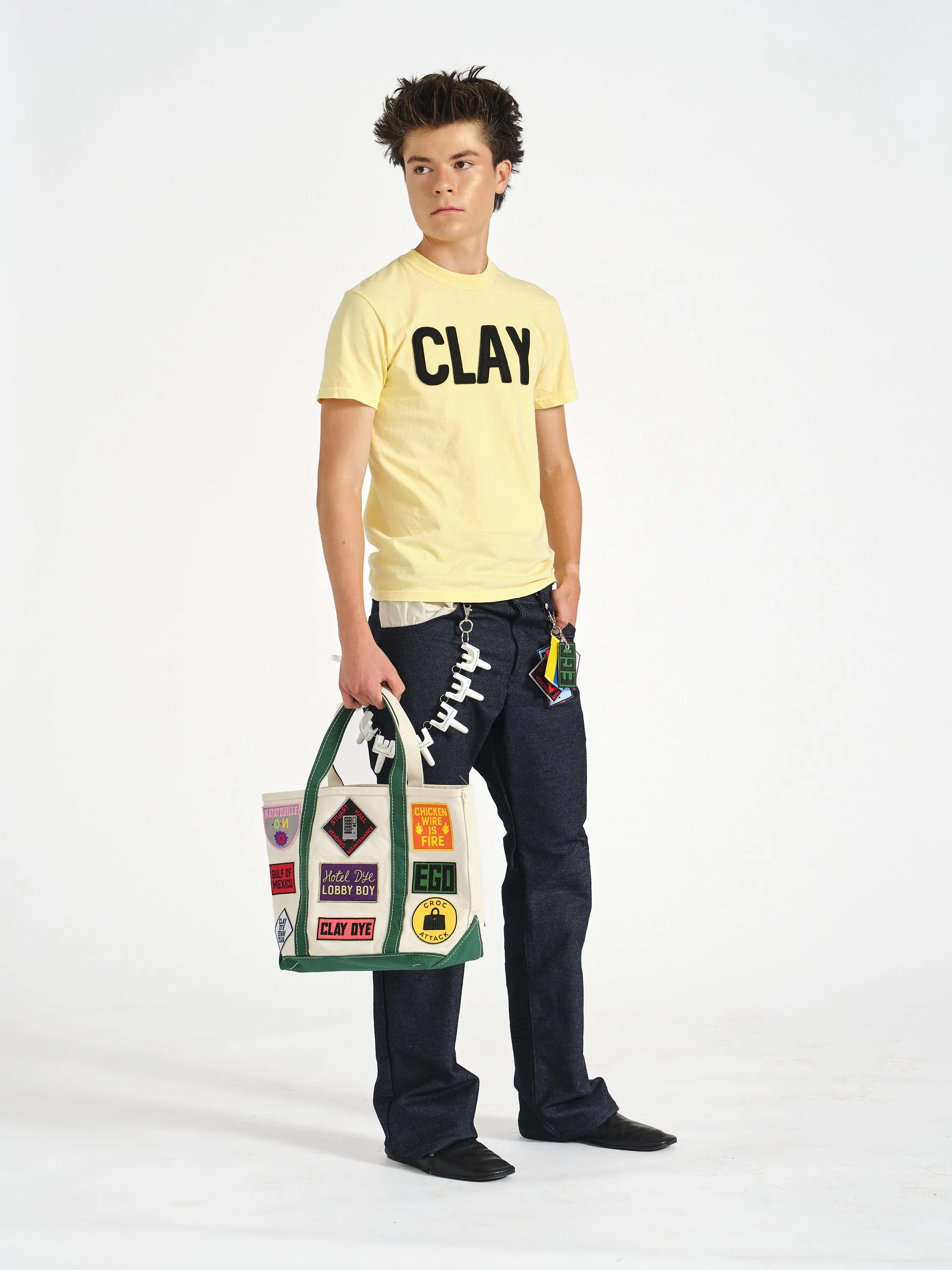 A male model with curly brown hair wearing a yellow T-shirt with the word 'CLAY' printed on it, dark jeans, black shoes, and holding a large tote bag with multiple colorful patches.