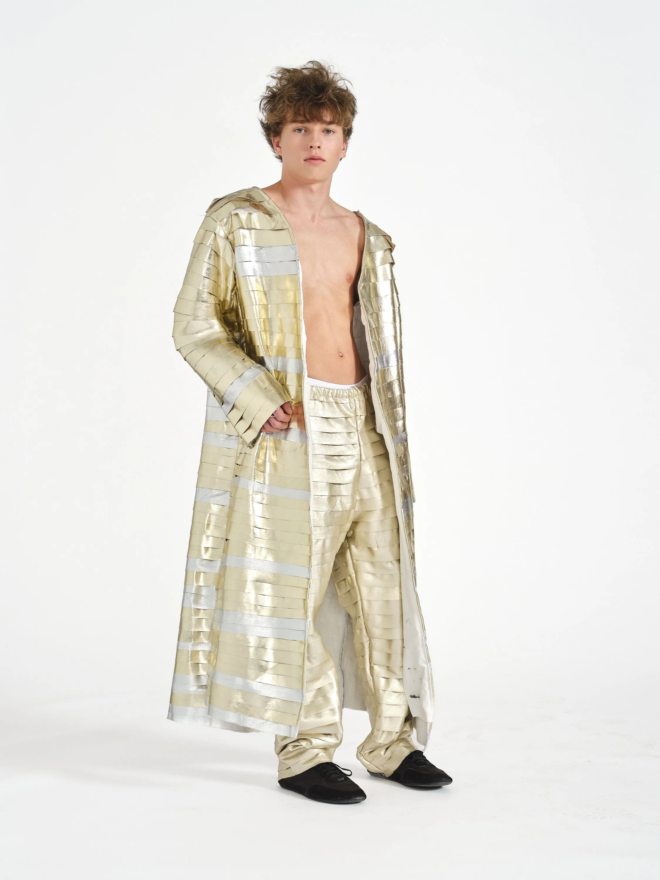 A male model standing in front of a plain white background, wearing beige and gold patterned clothing including a long coat and baggy pants, with one hand in his pocket, black shoes, and tousled brown hair.