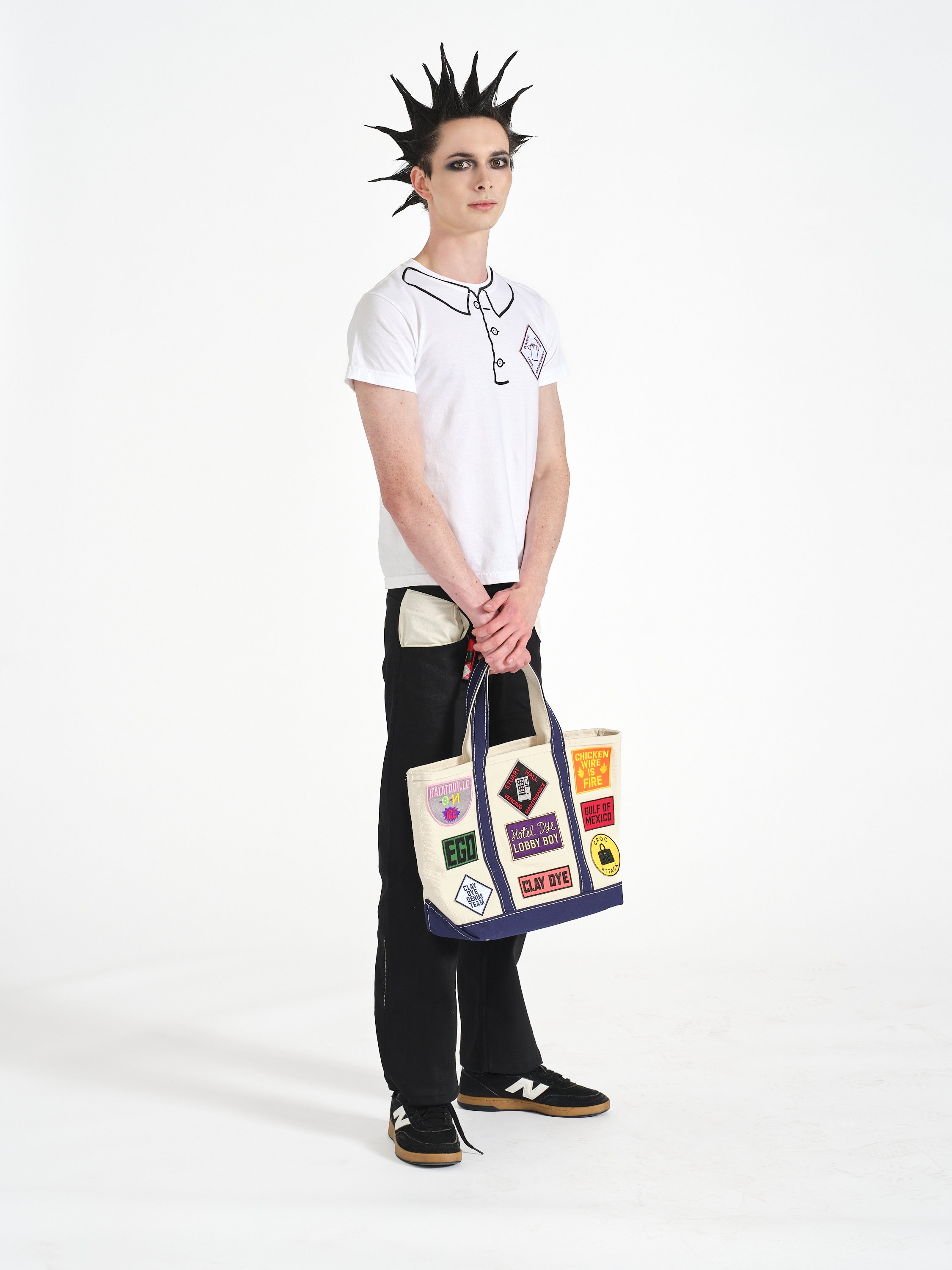 Model stands posed in Clay Dye denim, a white screenprinted t-shirt, and custom made canvas tote bag full of patches. He has a spiced mohawk and gothic makeup.