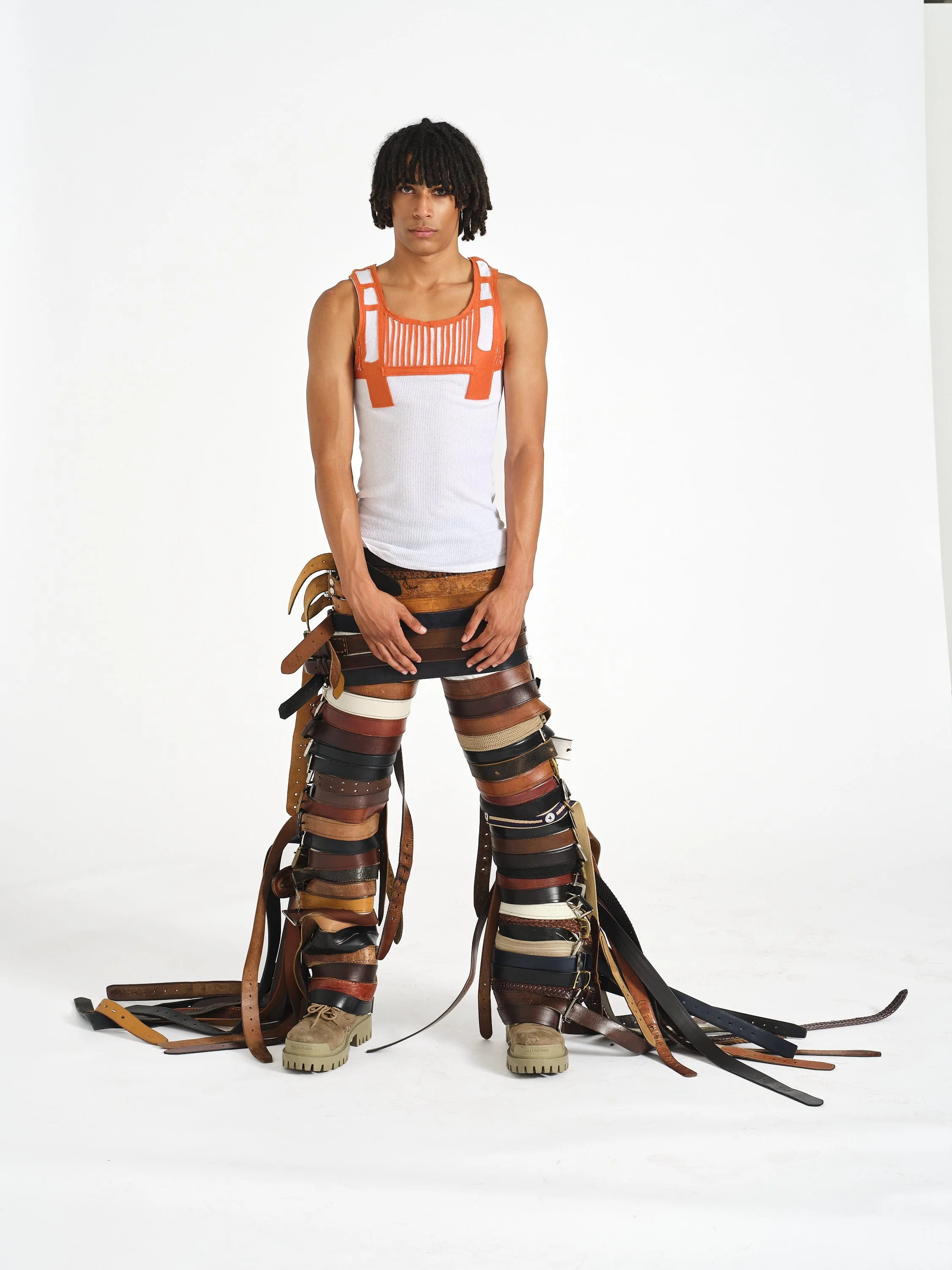 A male model with dreadlocks wearing a sleeveless top and tan boots standing against a plain white background. His pants are made from an array of belts and straps of various colors and widths that are draped over his legs and scattered on the floor.