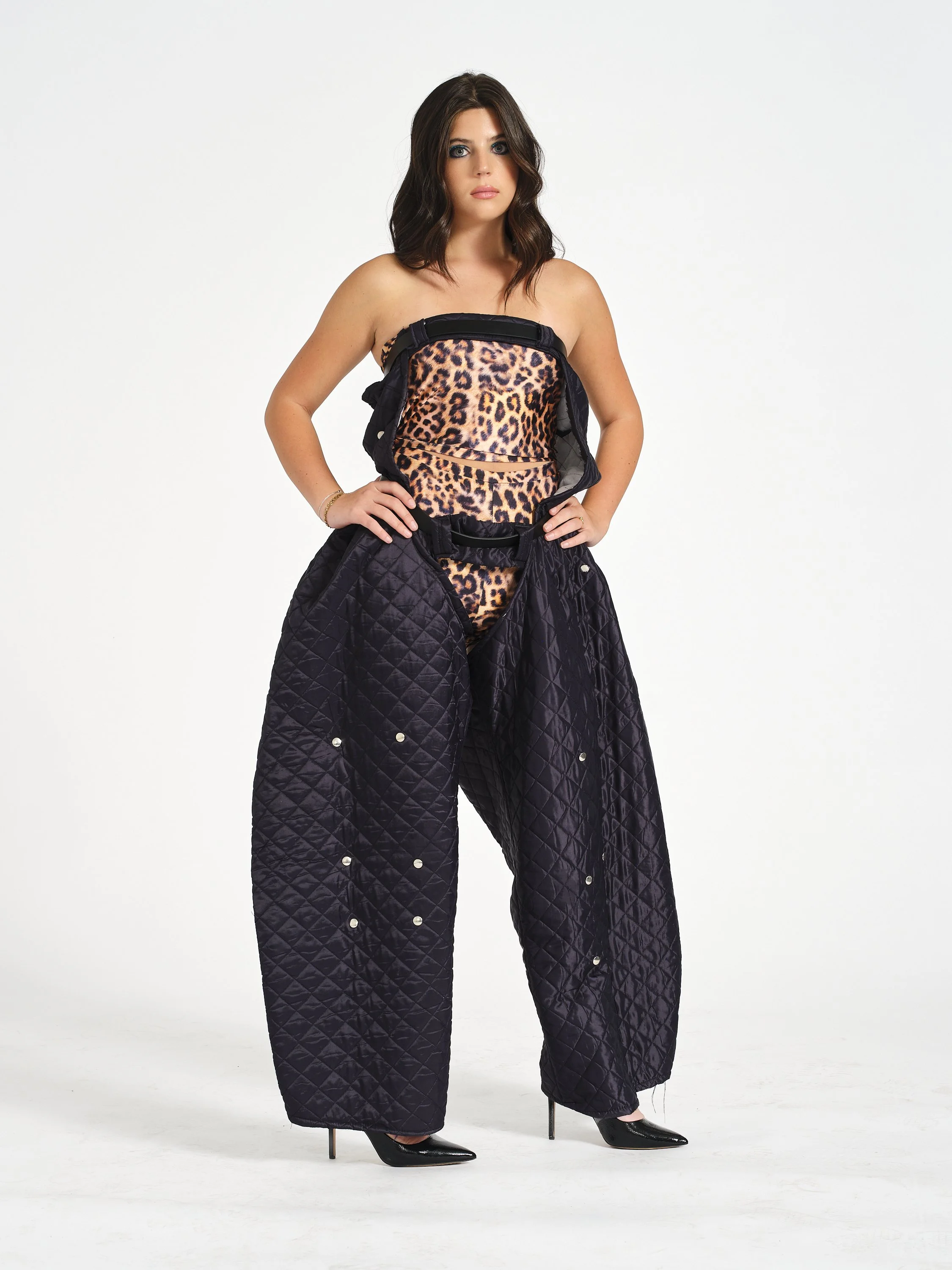 A female model wearing black high heels, a leopard print tube top, and black quilted quilted wide-leg chaps, standing with hands on hips against a white background.
