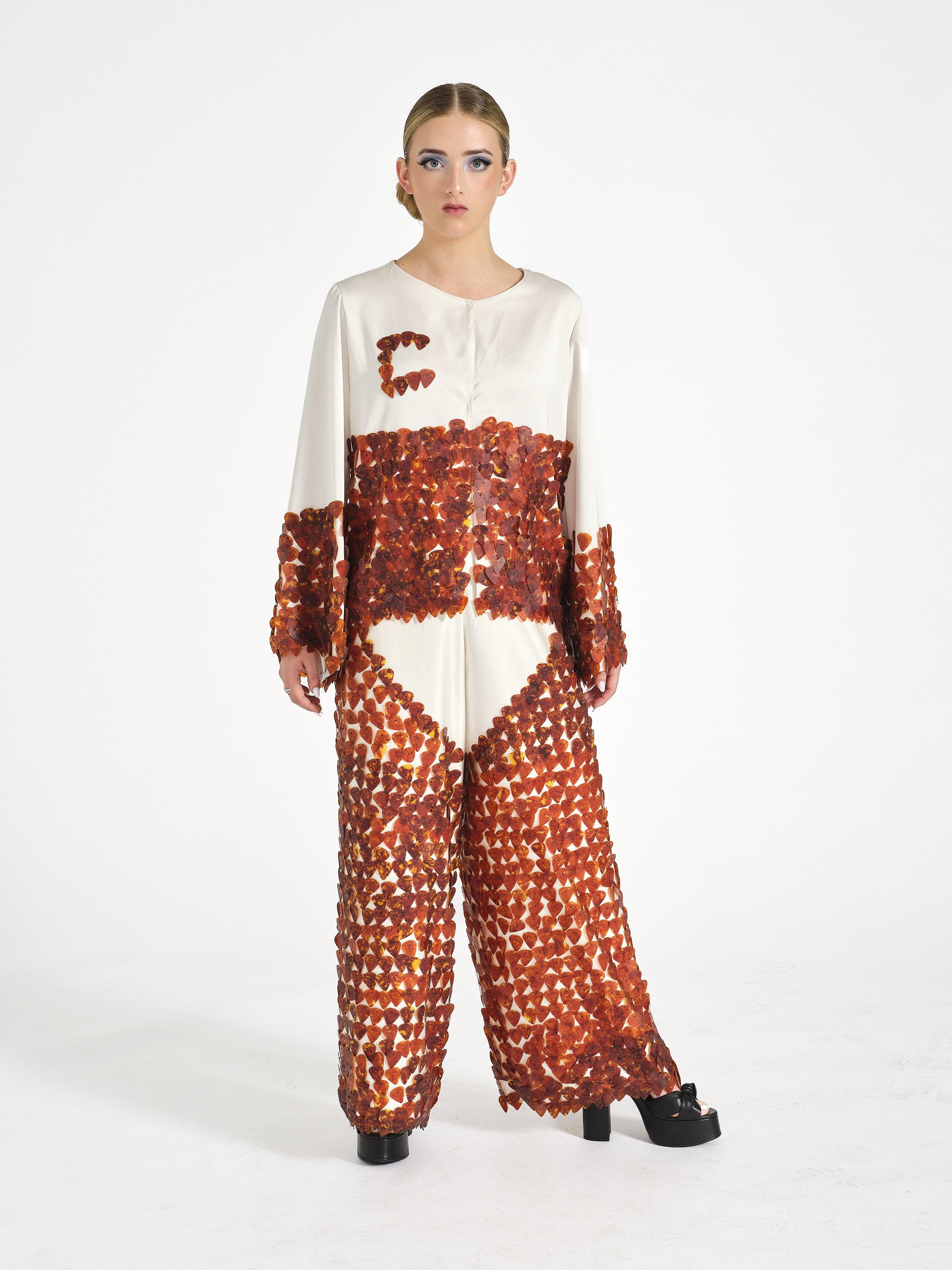A female model wearing a white jumpsuit decorated with numerous orange and brown guitar picks, standing against a plain white background.