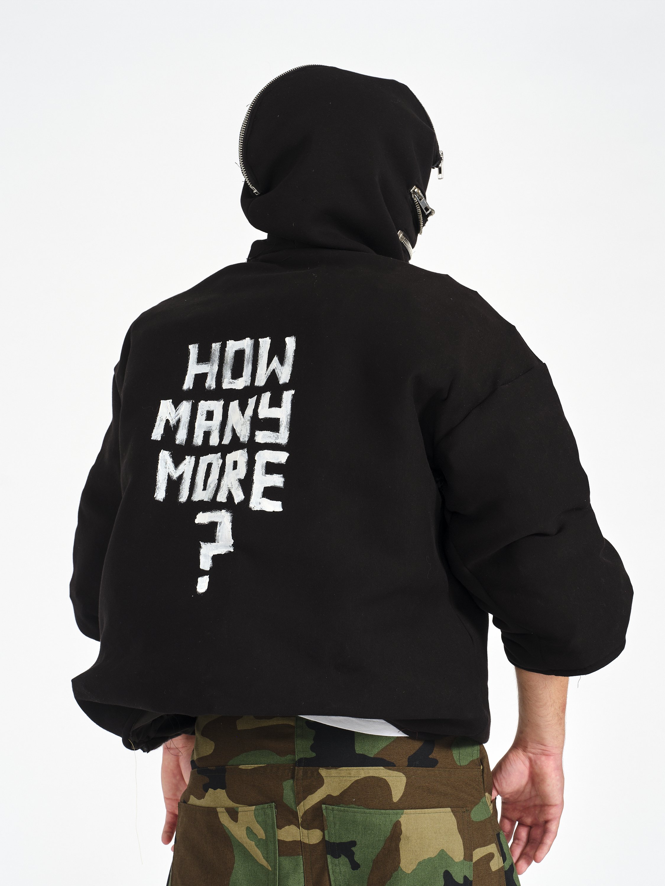 Back view of a person wearing a black zippered hoodie with white “HOW MANY MORE?” text and camouflage pants
