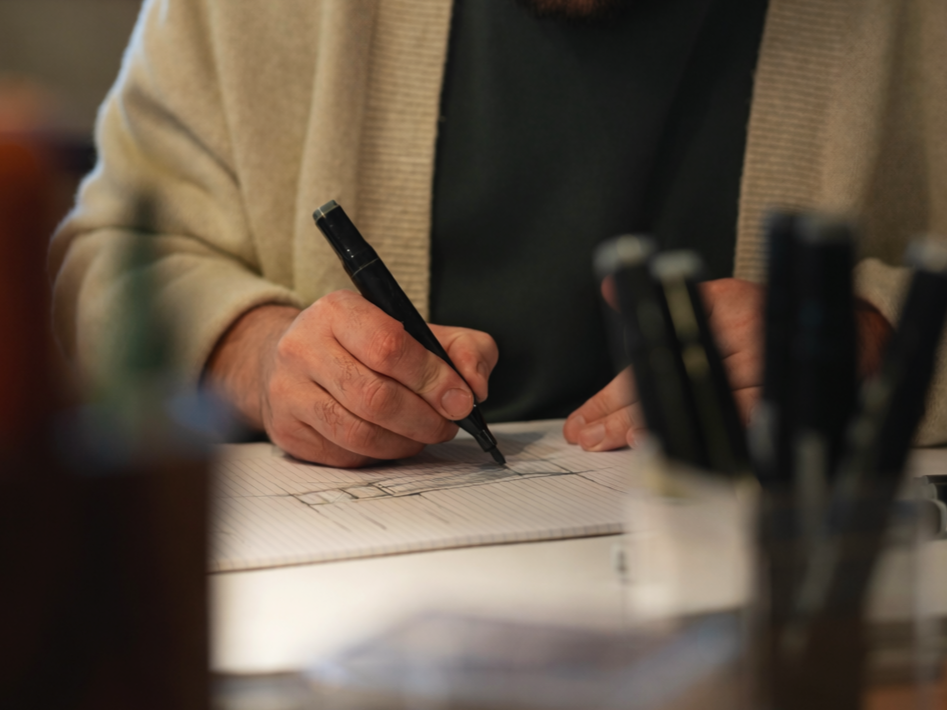Une personne était en train d'écrire ou de dessiner sur une feuille avec un stylo noir, avec un bloc-notes ou un carnet ouvert devant elle, dans un environnement intérieur.
