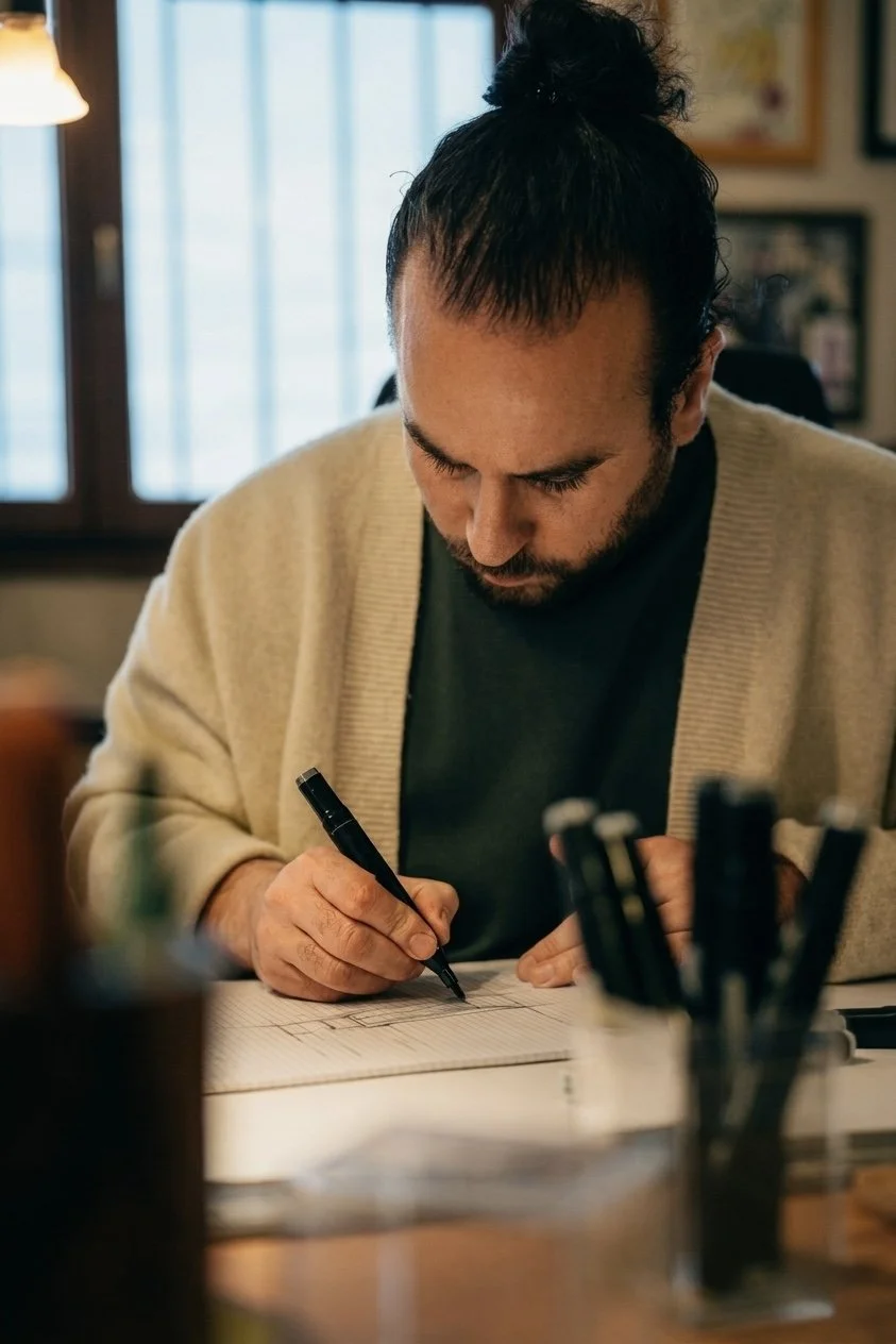 Un homme avec une barbe et un chignon, assis à un bureau, écrit sur un carnet avec un stylo. Il porte un pull beige et un t-shirt noir, dans une pièce éclairée par la lumière naturelle d'une fenêtre.