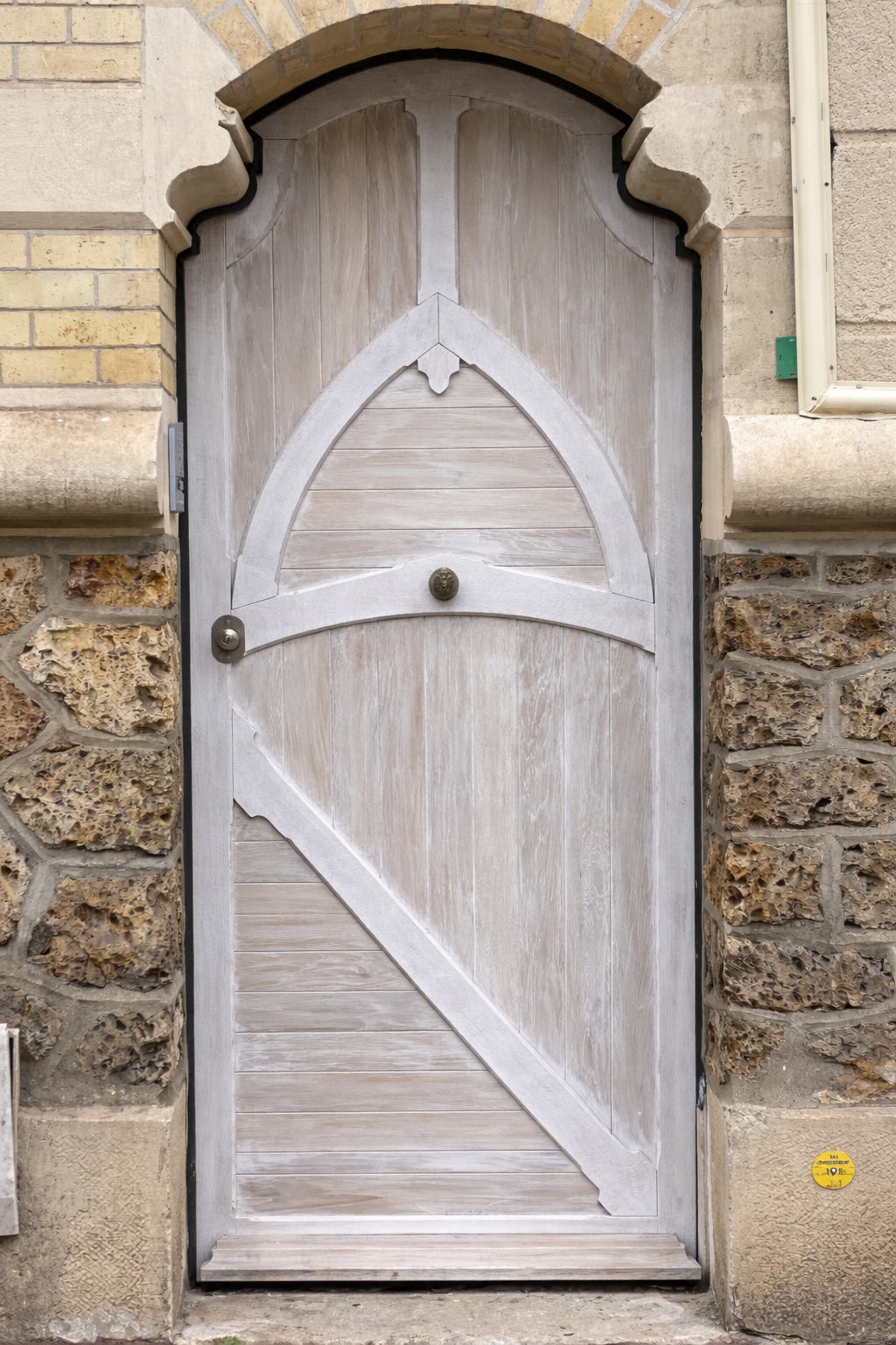 Porte en bois clair avec motif en forme d'arc, encadrée par des murs en pierre et briques, avec un bouton de porte en bronze.