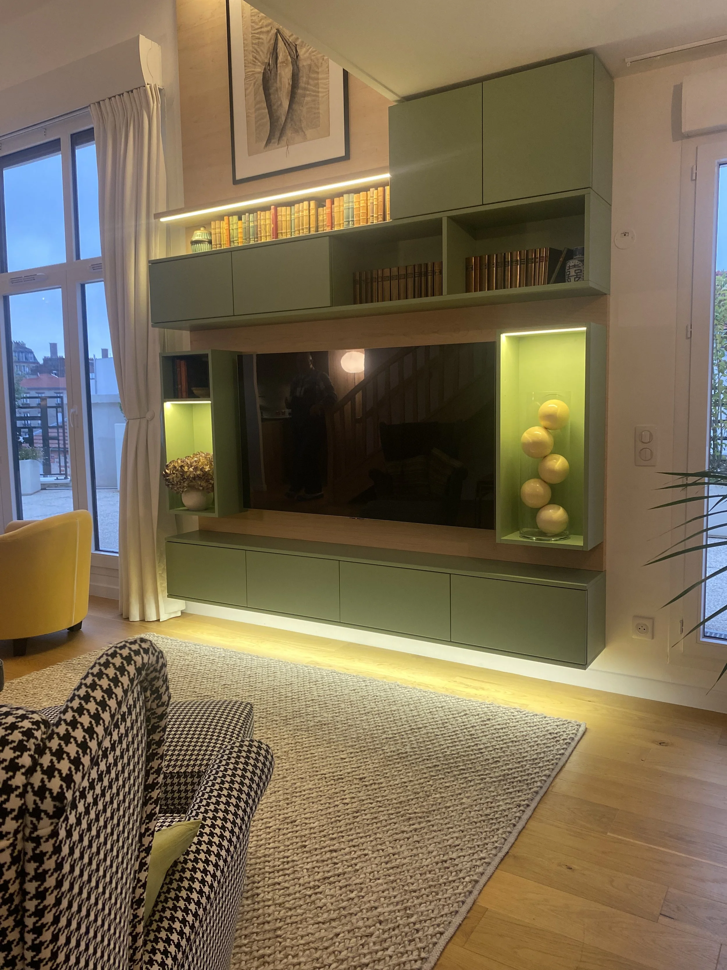 Salon avec mur de télévision, étagères vertes, plantes, fauteuils à motif pied-de-poules, tapis beige, grandes fenêtres avec rideaux, vue urbaine en soirée.
