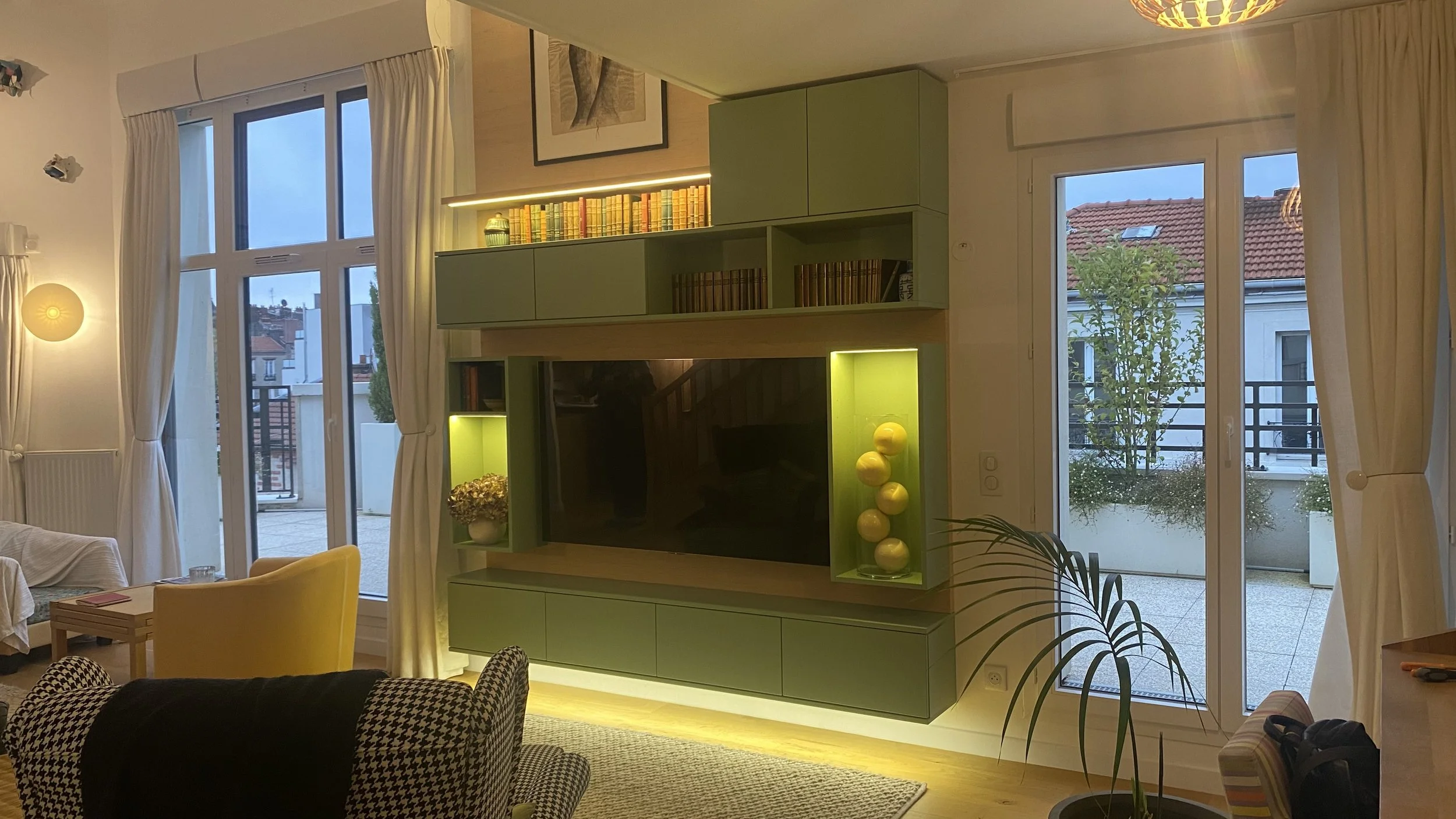 Intérieur d'un salon avec de grandes fenêtres, une télévision encadrée de placards verts, et des décorations modernes avec des livres et des lampes.