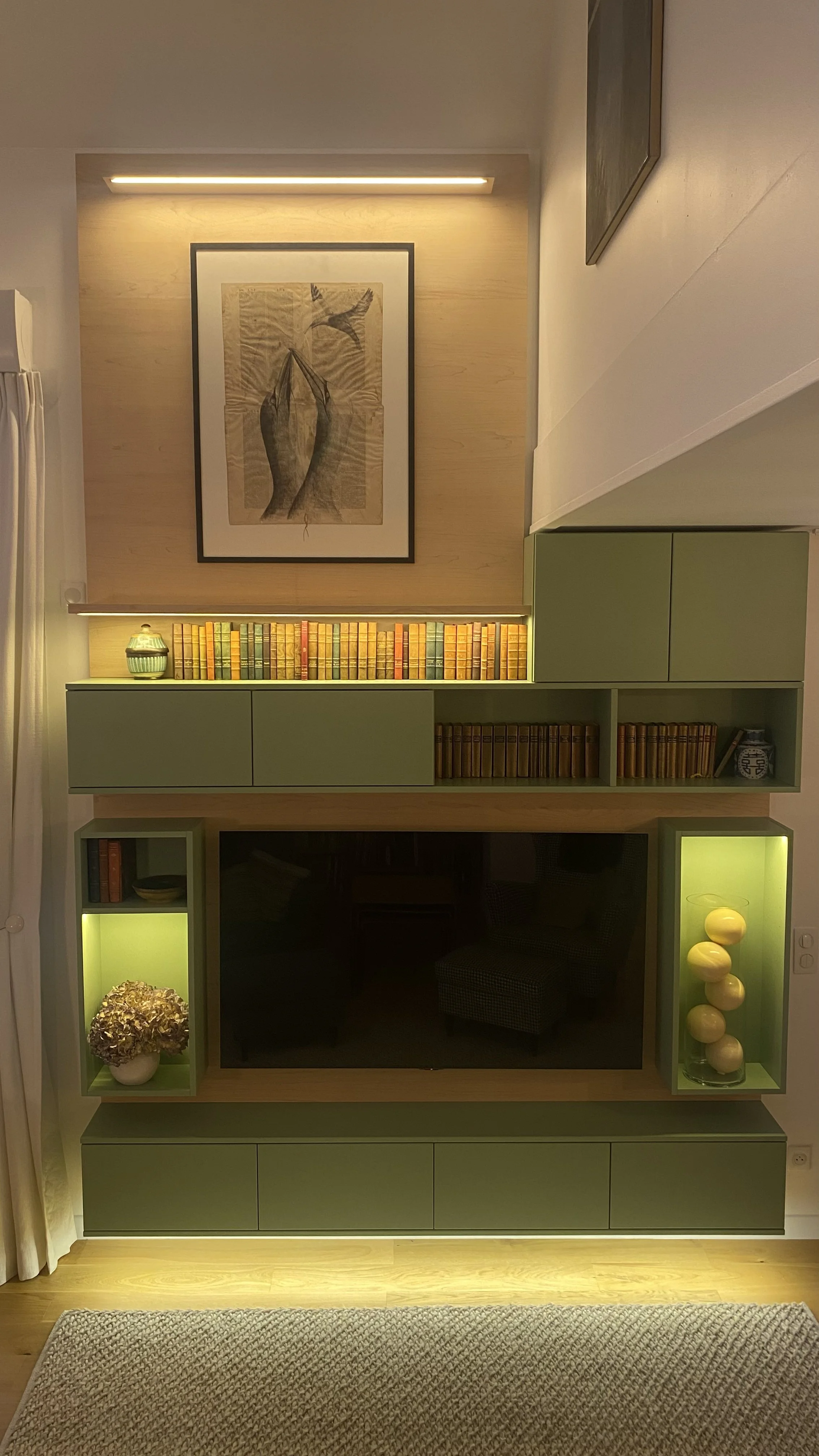 Une étagère murale verte contenant des livres, un vase, des décorations jaunes, une plante et une télévision, avec un tableau suspendu au-dessus et une lumière LED.