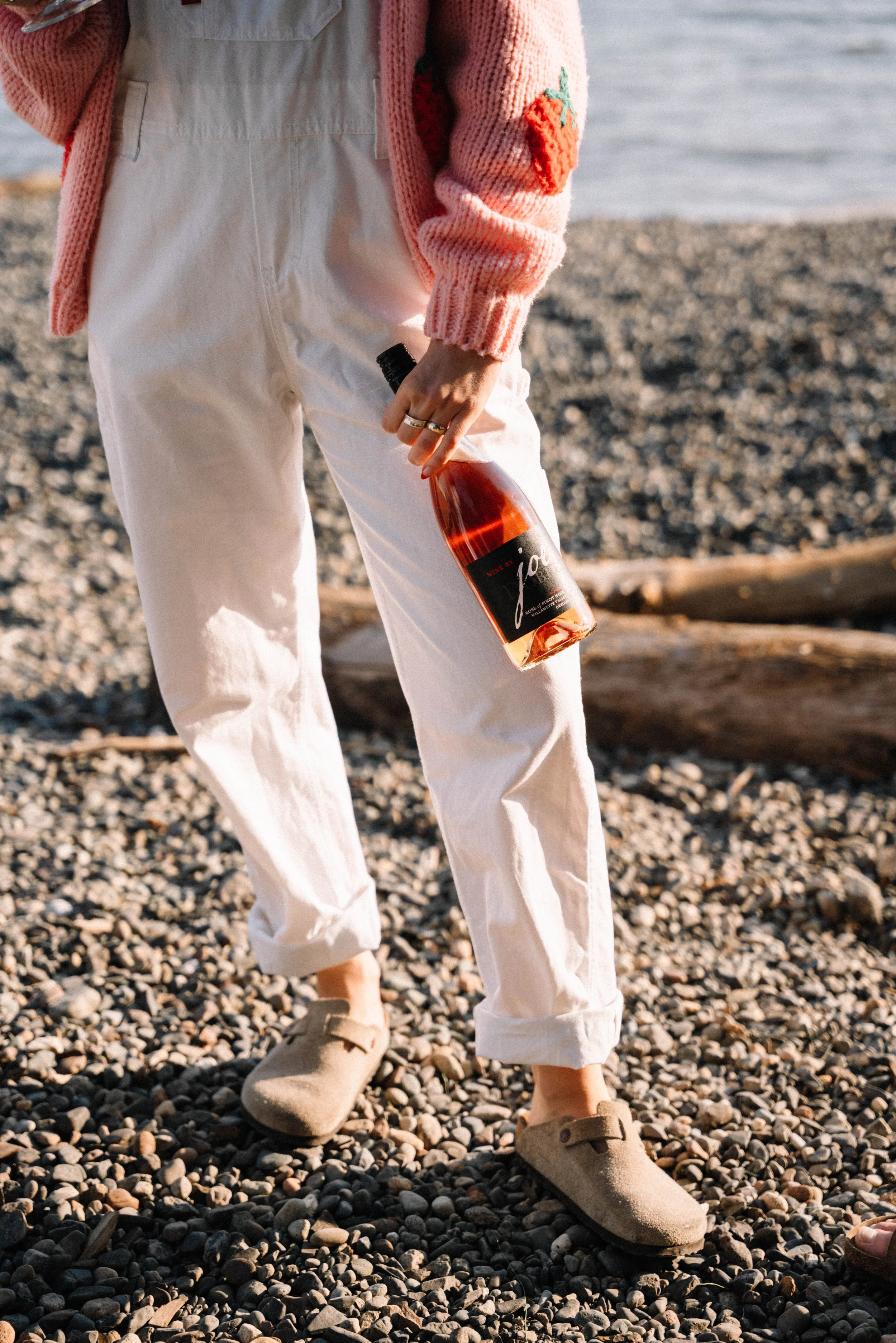 Person standing on a pebble beach, holding a bottle of rosé wine, wearing beige slippers, white pants, and a pink sweater with a red flower patch.