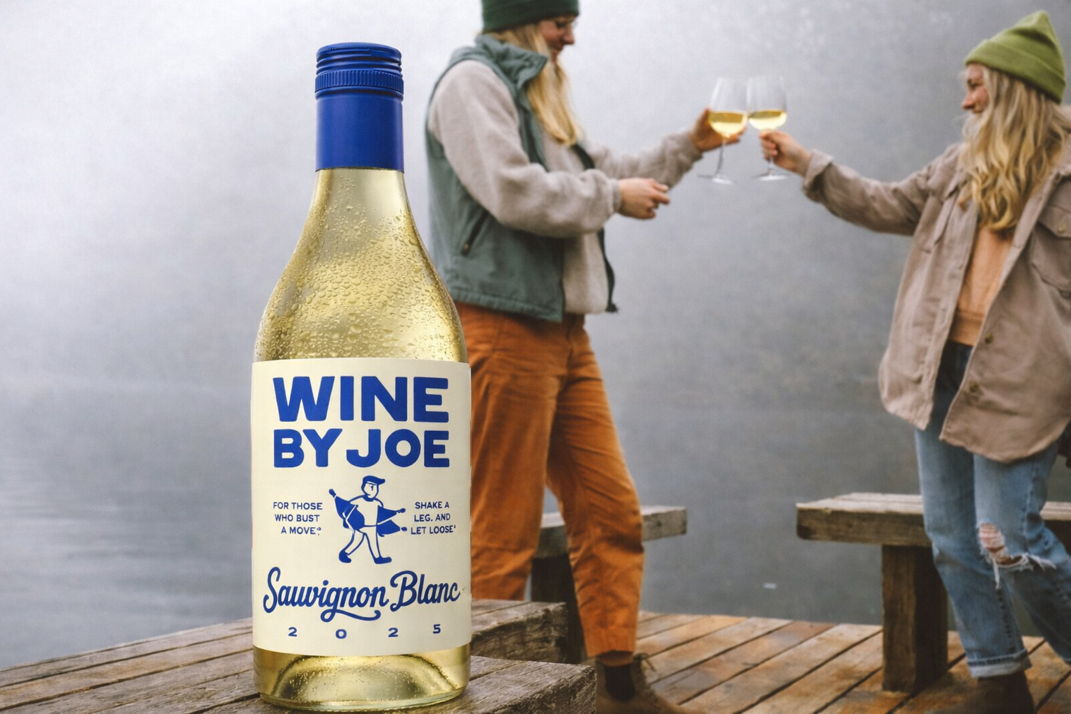 A bottle of wine labeled 'WINE BY JOE, Sauvignon Blanc 2025' in the foreground, with two women in casual outdoor clothing raising glasses of white wine in a toast on a wooden dock by a lake in the background.