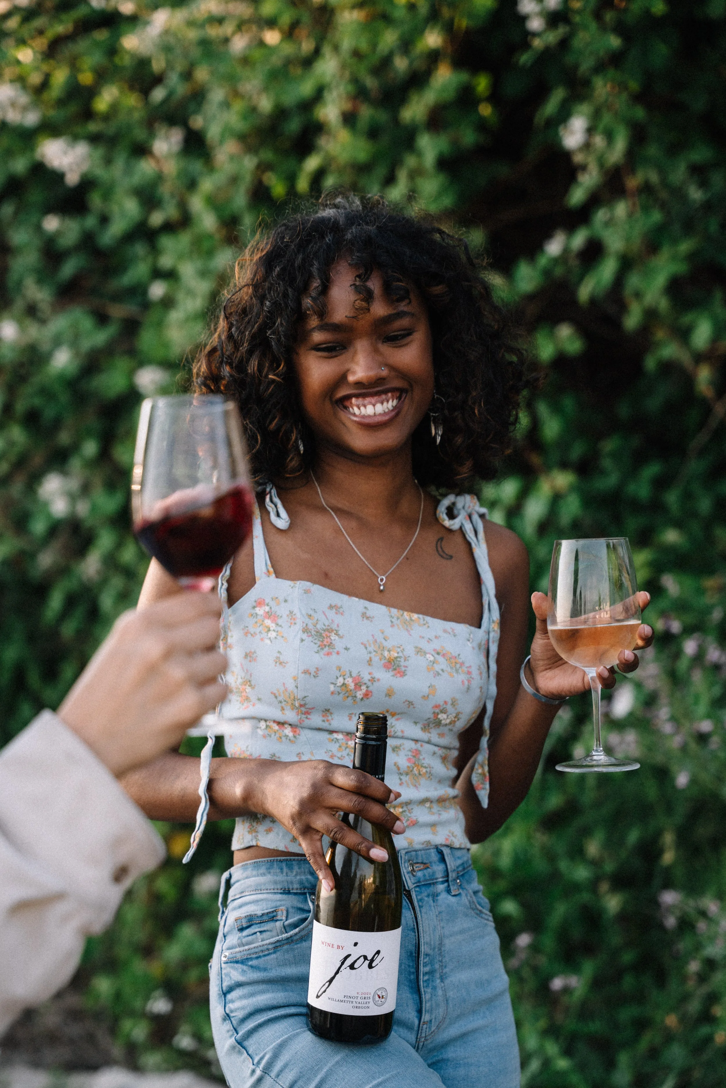 A young woman with curly hair smiling outdoors, holding a glass of rosé wine in one hand and a wine bottle in the other, with another person partially visible holding a glass of red wine.